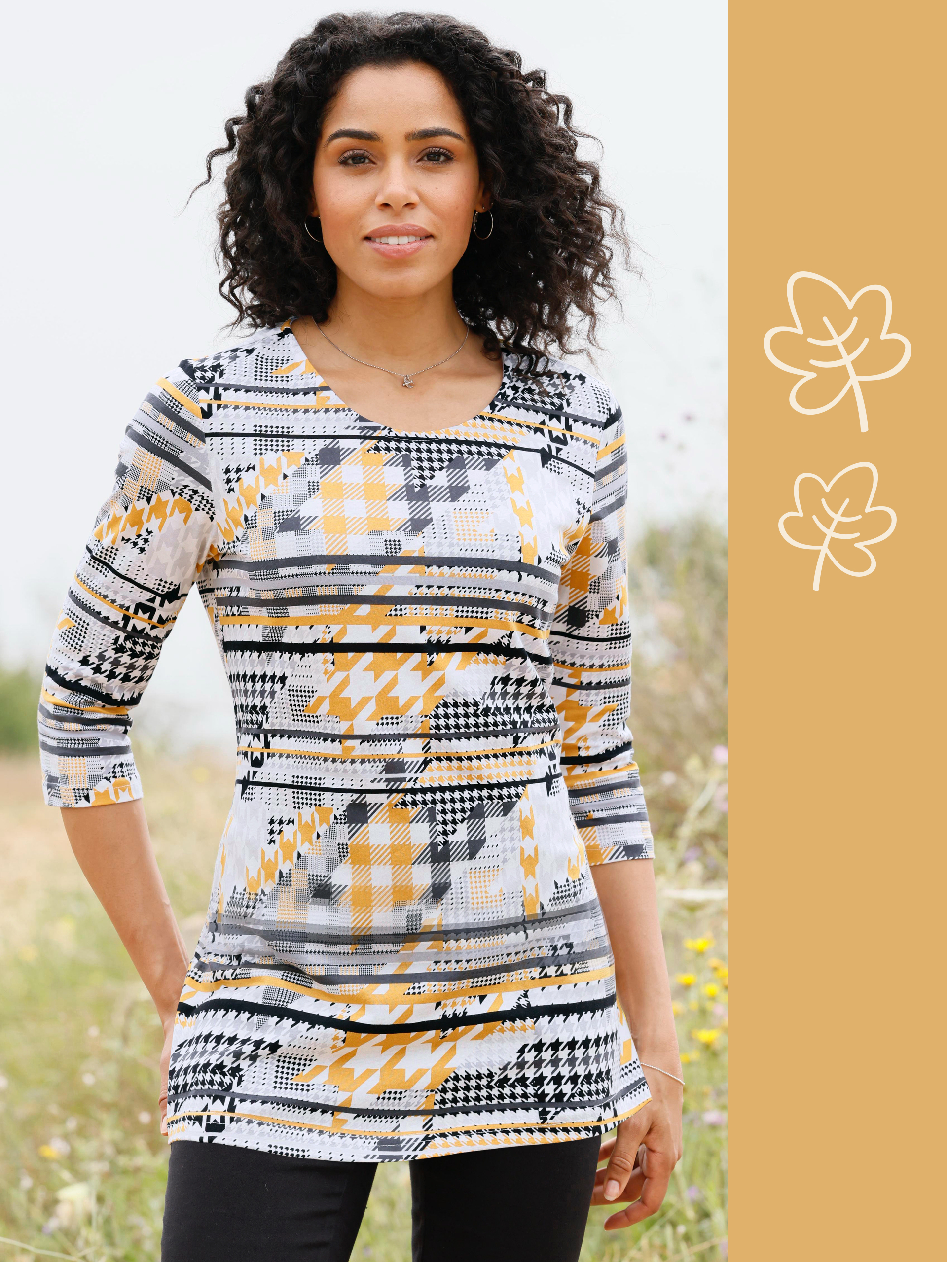 En modell bär en mönstrad topp i gult, svart och vitt. Den står i en blomstrande, naturlig miljö.