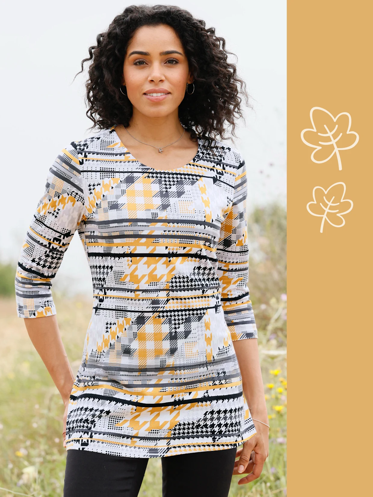 Ein Model trägt ein gemustertes Oberteil in Gelb, Schwarz und Weiß. Es steht in einer blühenden, natürlichen Umgebung.