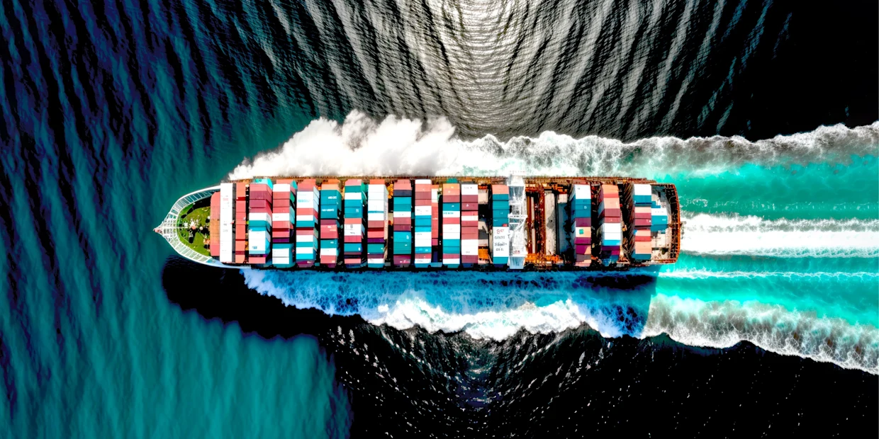 Luftaufnahme eines Containerschiffs mit bunten Frachtcontainern, das durch türkisfarbenes Wasser fährt und weiße Gischt hinterlässt. Luftaufnahme eines Containerschiffs mit bunten Frachtcontainern, das durch türkisfarbenes Wasser fährt und weiße Gischt hinterlässt.