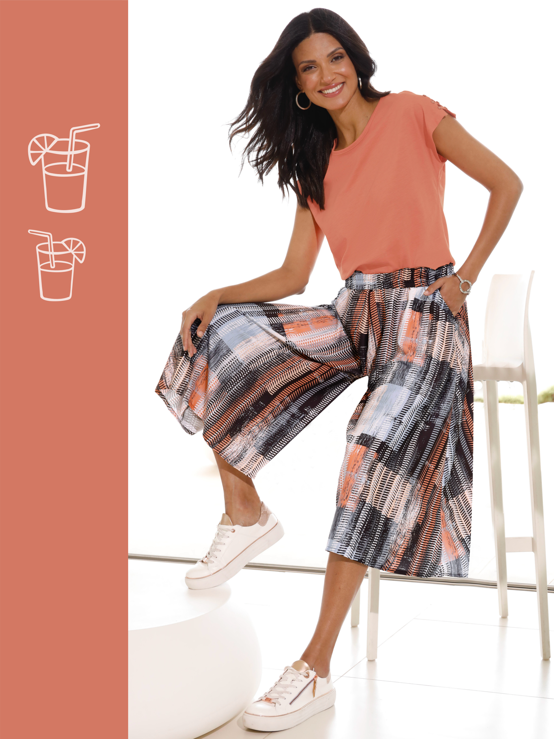 Frau trägt ein korallfarbenes T-Shirt, gemusterte Culotte-Hose in Schwarz, Weiß und Koralltönen sowie weiße Sneaker mit goldenen Details.