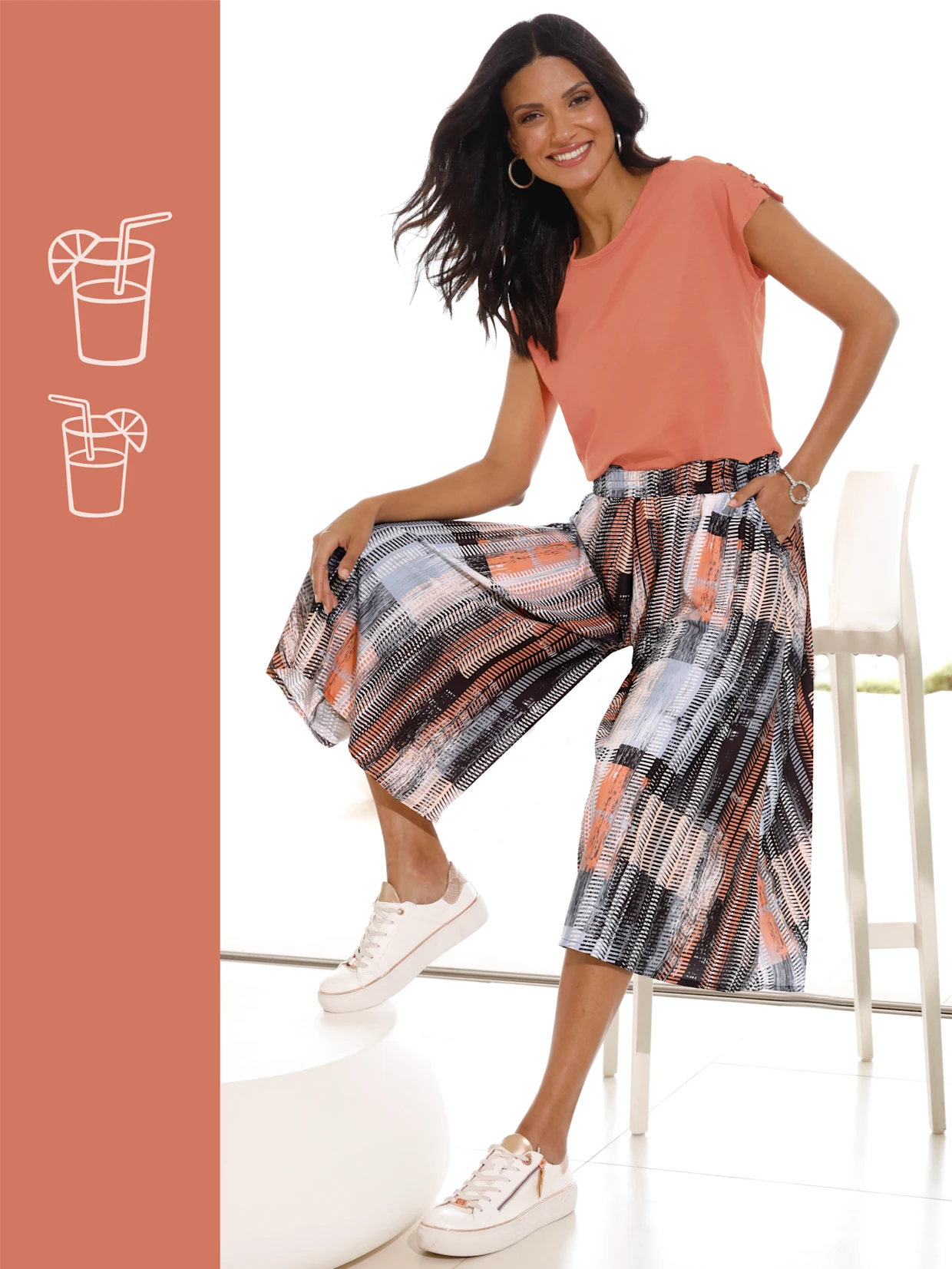 Frau trägt ein korallfarbenes T-Shirt, gemusterte Culotte-Hose in Schwarz, Weiß und Koralltönen sowie weiße Sneaker mit goldenen Details. Frau trägt ein korallfarbenes T-Shirt, gemusterte Culotte-Hose in Schwarz, Weiß und Koralltönen sowie weiße Sneaker mit goldenen Details.