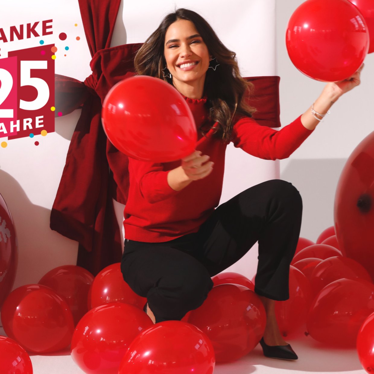 Frau mit rotem Oberteil vor Geschenkpaket mit roten Luftballons in der Hand