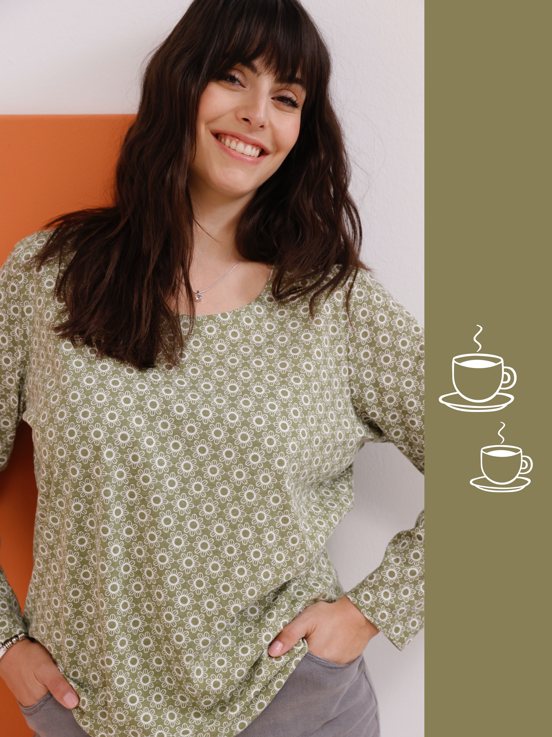 Ein Model steht in einem blumigen, olivgrünen Oberteil und grauen Hosen, vor einem orangefarbenen Hintergrund.