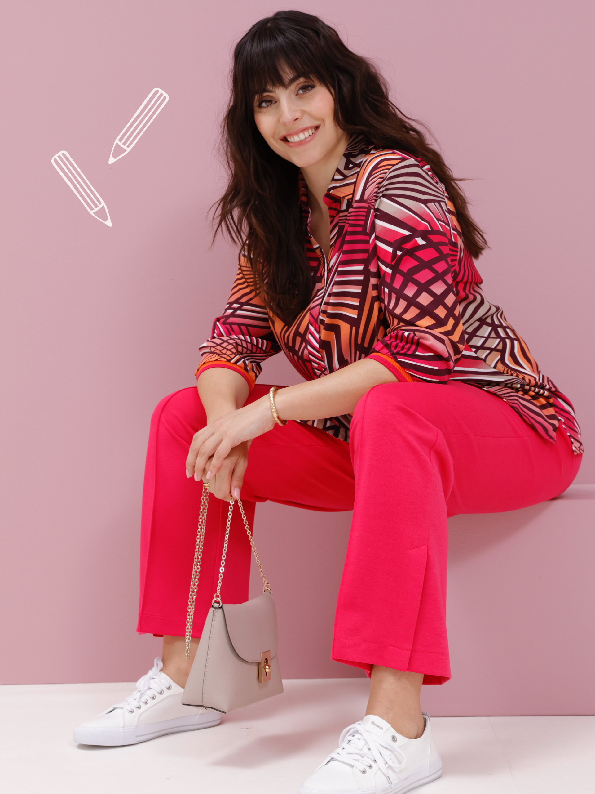 Ein Model sitzt vor einem rosa Hintergrund und präsentiert eine farbenfrohe, gemusterte Bluse, eine pinke Hose, weiße Sneaker und eine Handtasche.