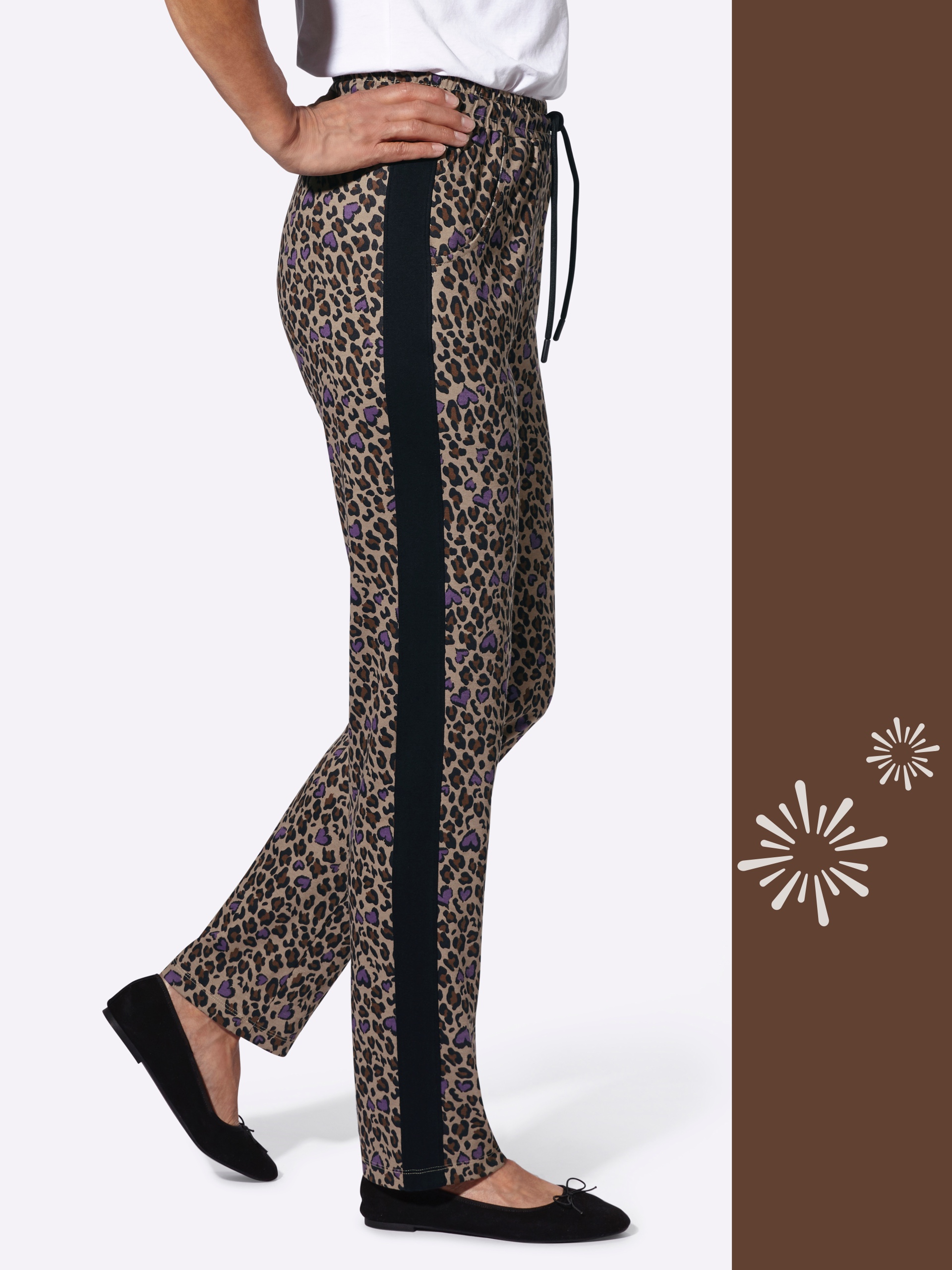 Ein Model trägt eine gemusterte Hose mit seitlichem schwarzen Streifen und schwarzen Ballerinas, Hände auf der Hüfte.