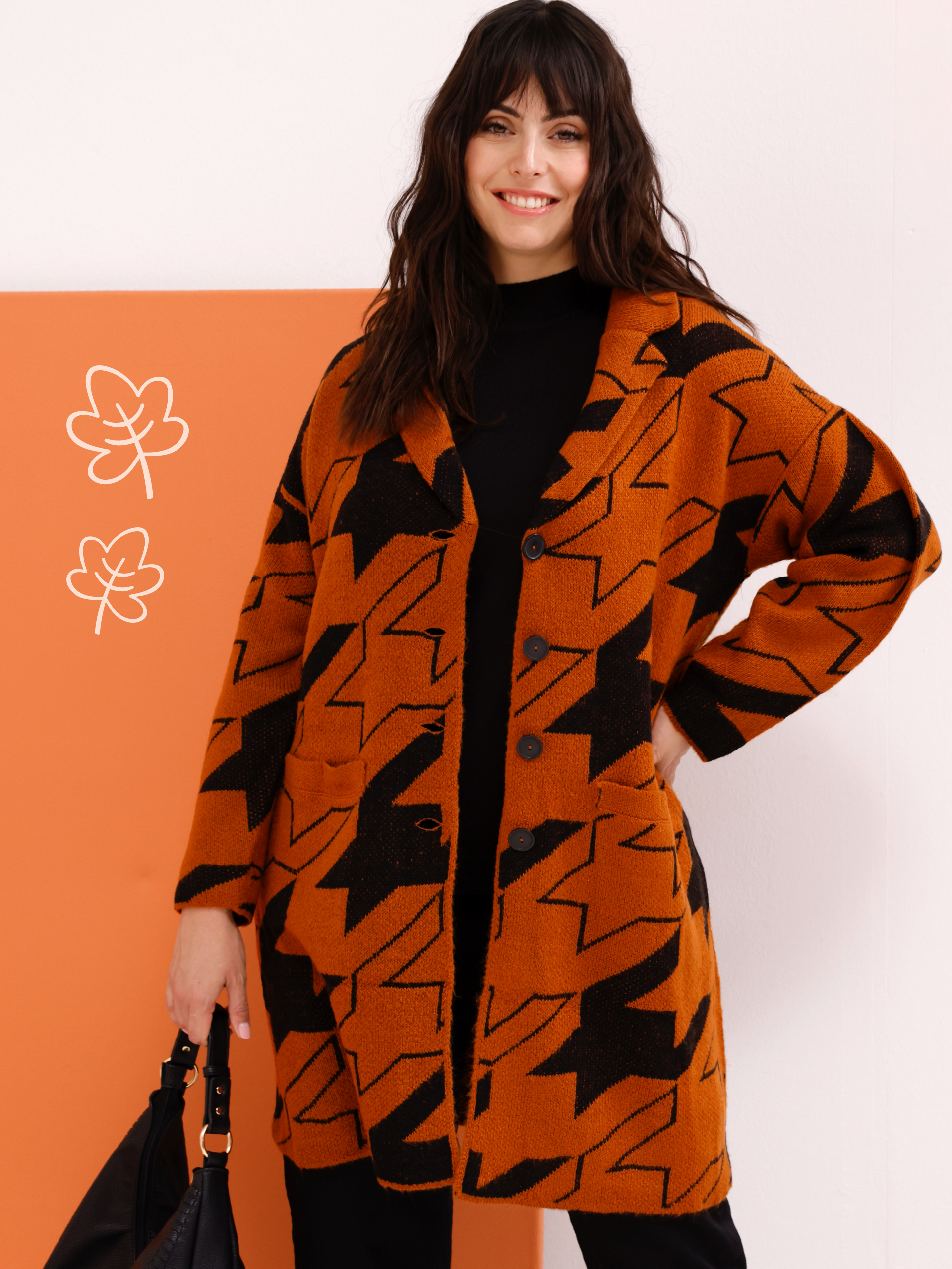 Ein Model trägt eine lange, orange-schwarze Strickjacke mit großem Muster über einem schwarzen Oberteil, dazu hält es eine schwarze Tasche.
