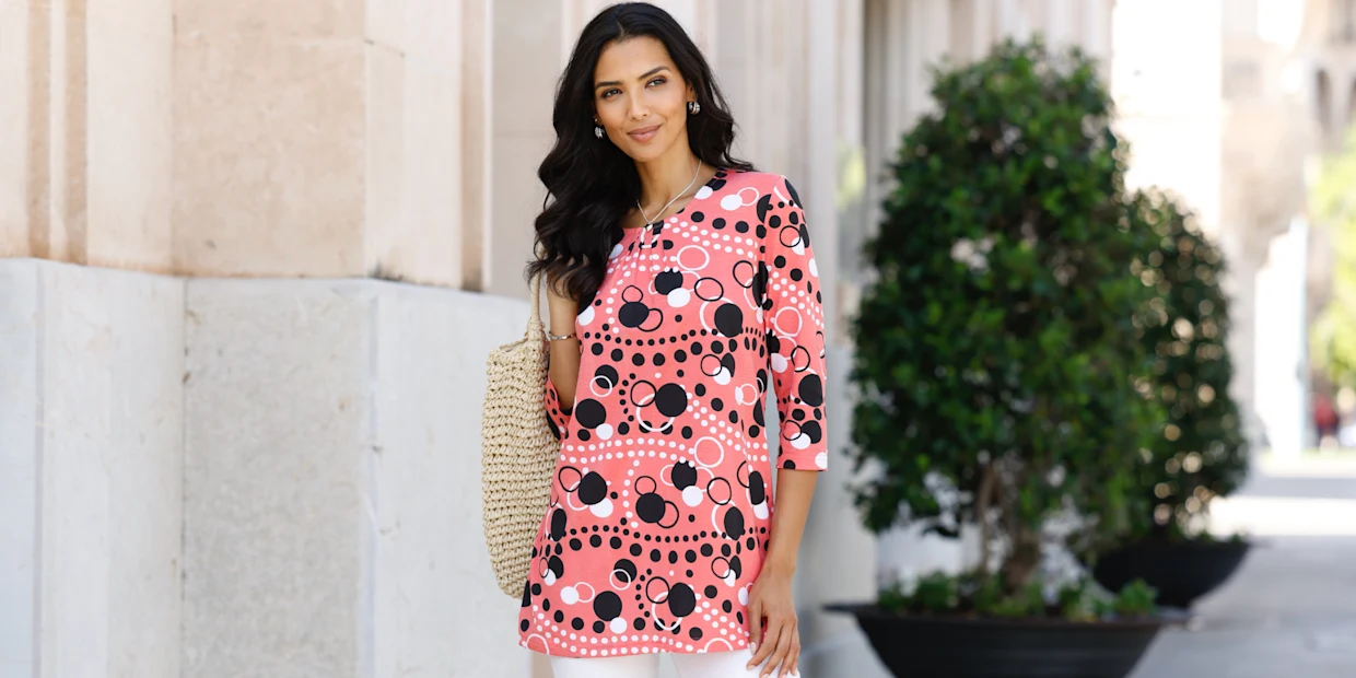 Model má na sobě růžové puntíkované tričko s 3/4 rukávy a drží pletenou tašku.