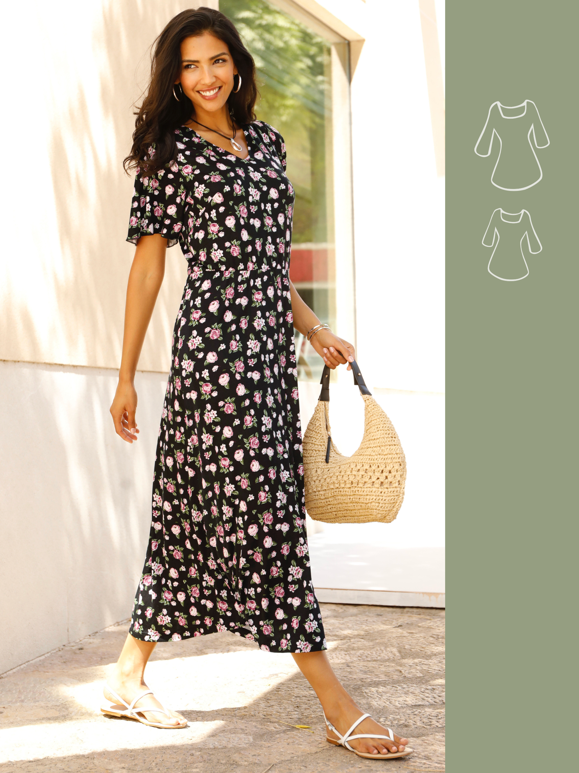 Ein Model trägt ein schwarzes Maxikleid mit rosa Blumenmuster, weiße Sandalen und hält eine geflochtene Tasche, steht im Sonnenlicht.