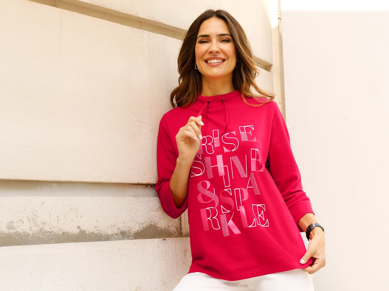 Modelka prezentuje ružovú mikinu s kapucňou s nápisom 'Rise Shine & Sparkle' a má na sebe biele nohavice.