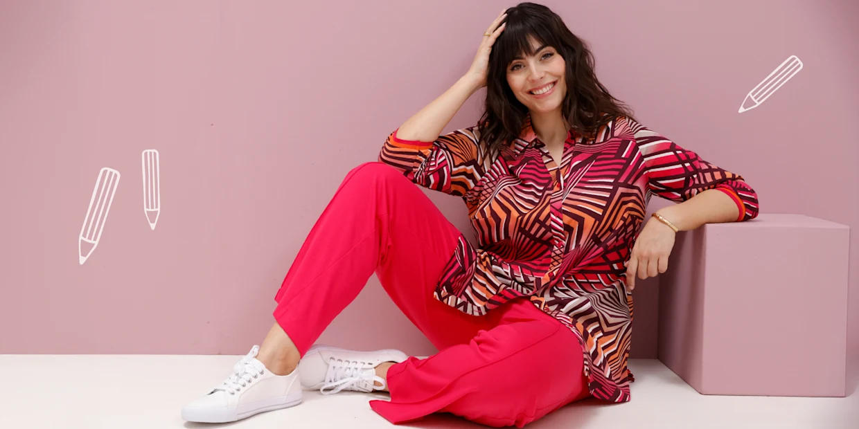 Ein Model sitzt in einer bunten Bluse und roten Hose vor einer rosa Wand. Ein Model sitzt in einer bunten Bluse und roten Hose vor einer rosa Wand.