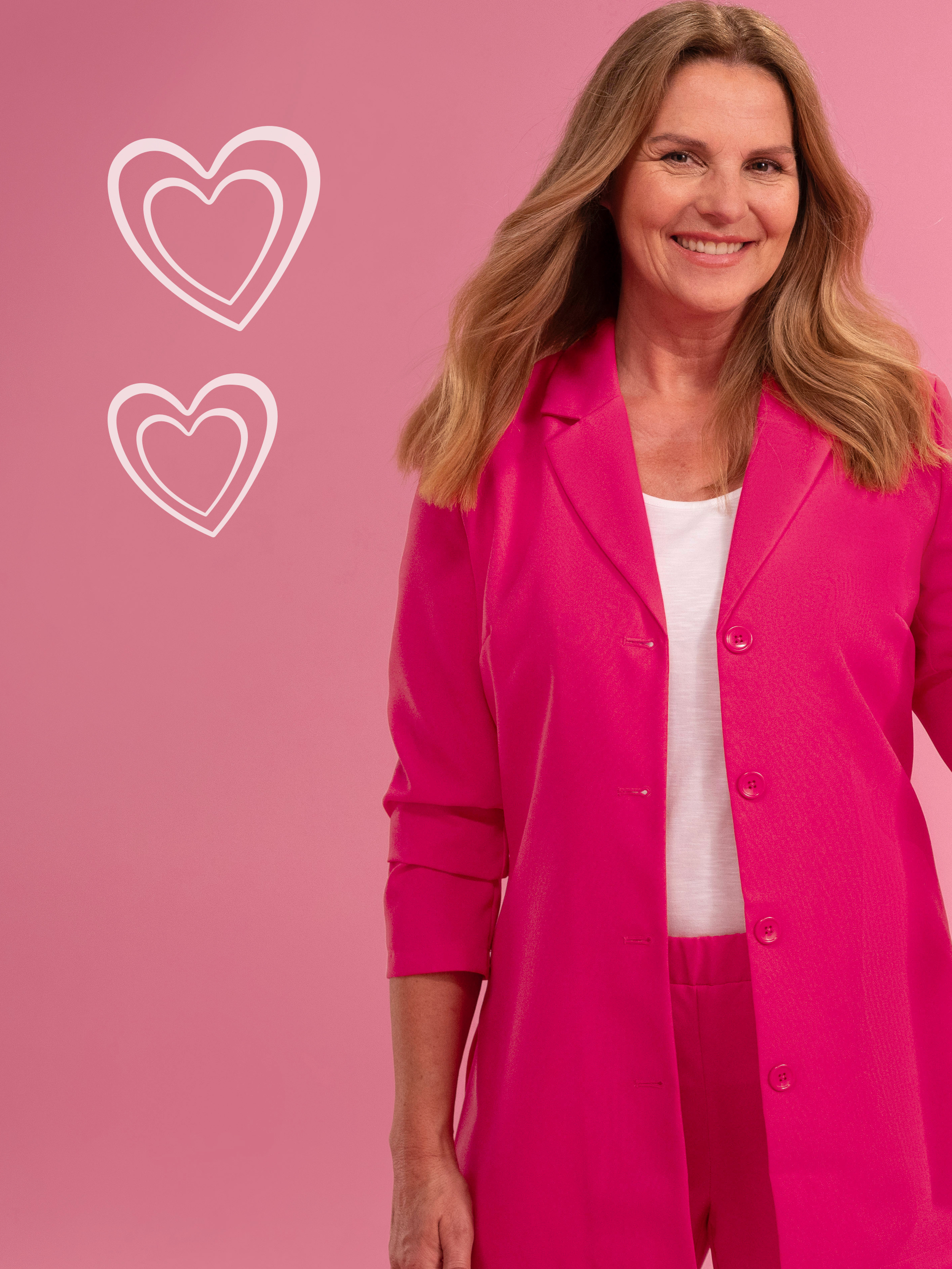 Frau trägt ein leuchtend pinkes Outfit vor einem rosa Hintergrund, bestehend aus einem Blazer und passender Hose; daneben zwei gezeichnete Herzen.