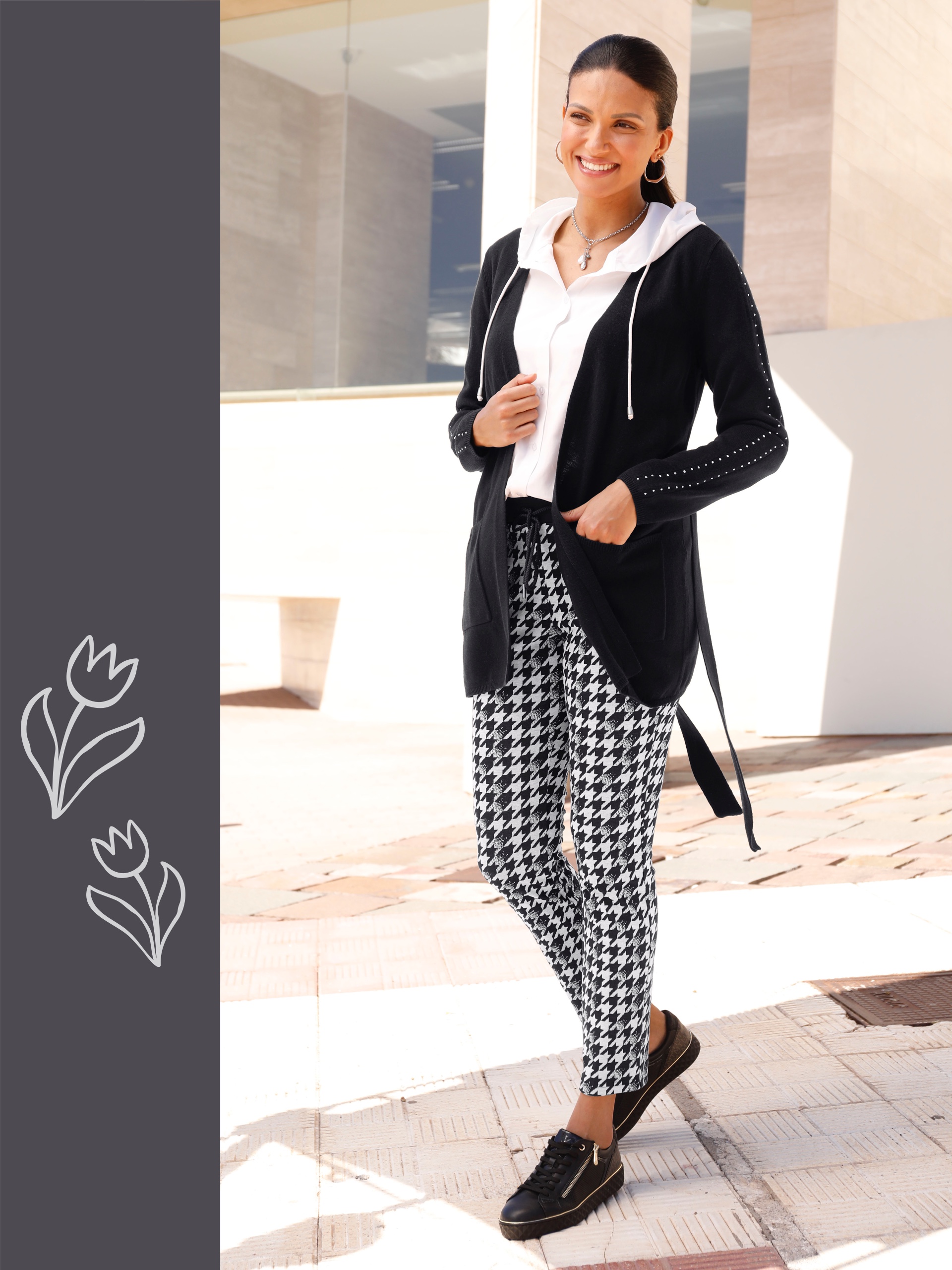 Ein Model trägt eine schwarze Jacke über einem weißen Oberteil und gemusterte Hosen. Der Hintergrund ist hell und modern.