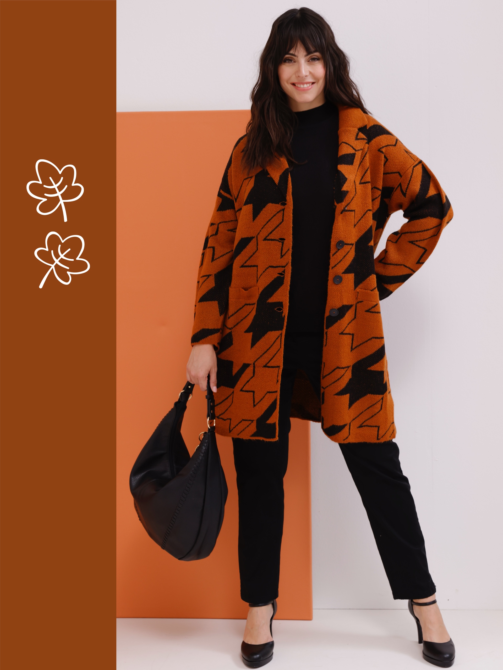 Ein Model trägt einen orange-schwarzen Cardigan und schwarze Hosen, während es eine schwarze Tasche hält, vor einem orangefarbenen Hintergrund.

