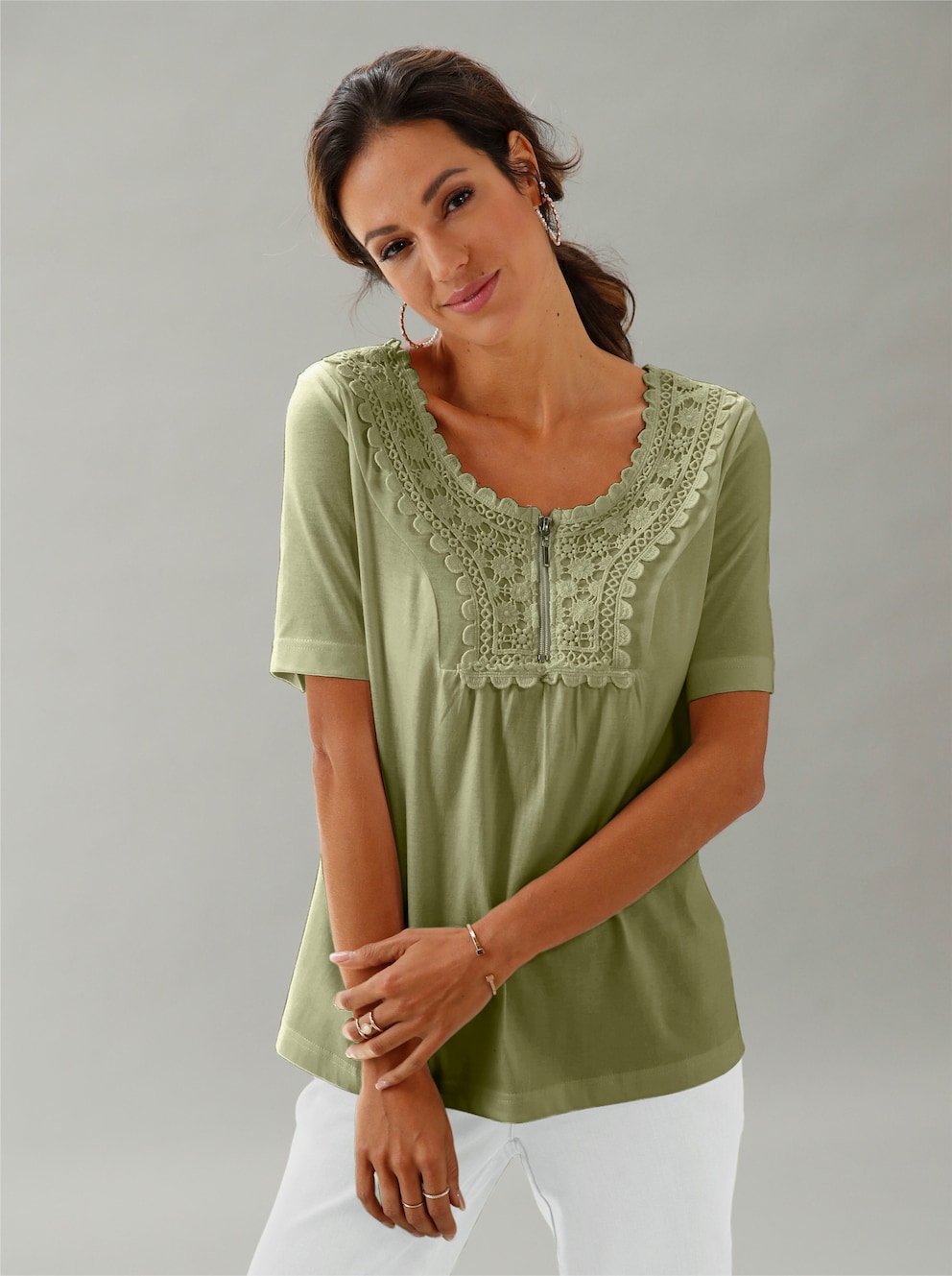 Frau mit grünem Kurzarm-Shirt mit wunderschöner Ton-in-Ton Spitze im Vorderteil. Mit Rundhals-Ausschnitt und kurzem Reißverschluss. Dazu trägt sie eine weiße Hose.