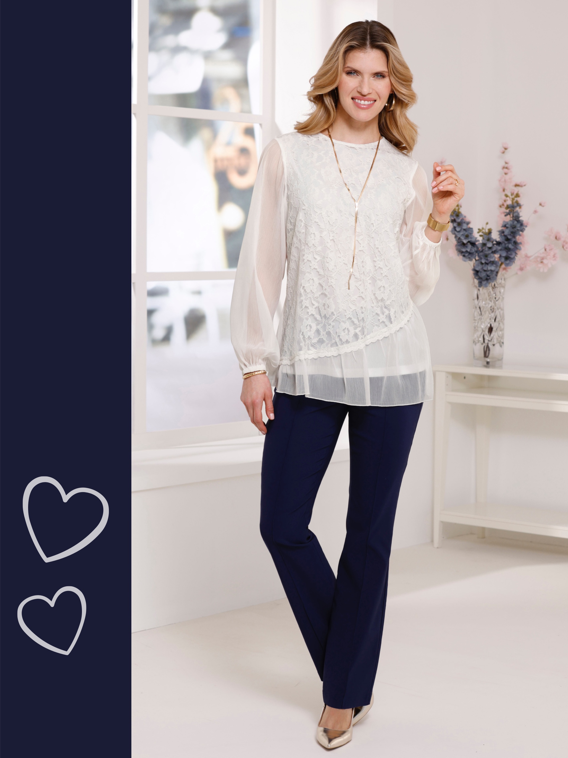 Ein Model trägt eine weiße, langärmelige Bluse mit Spitzenmuster und transparentem Saum sowie eine dunkelblaue Hose. Sie trägt goldfarbene Schuhe und Schmuck.