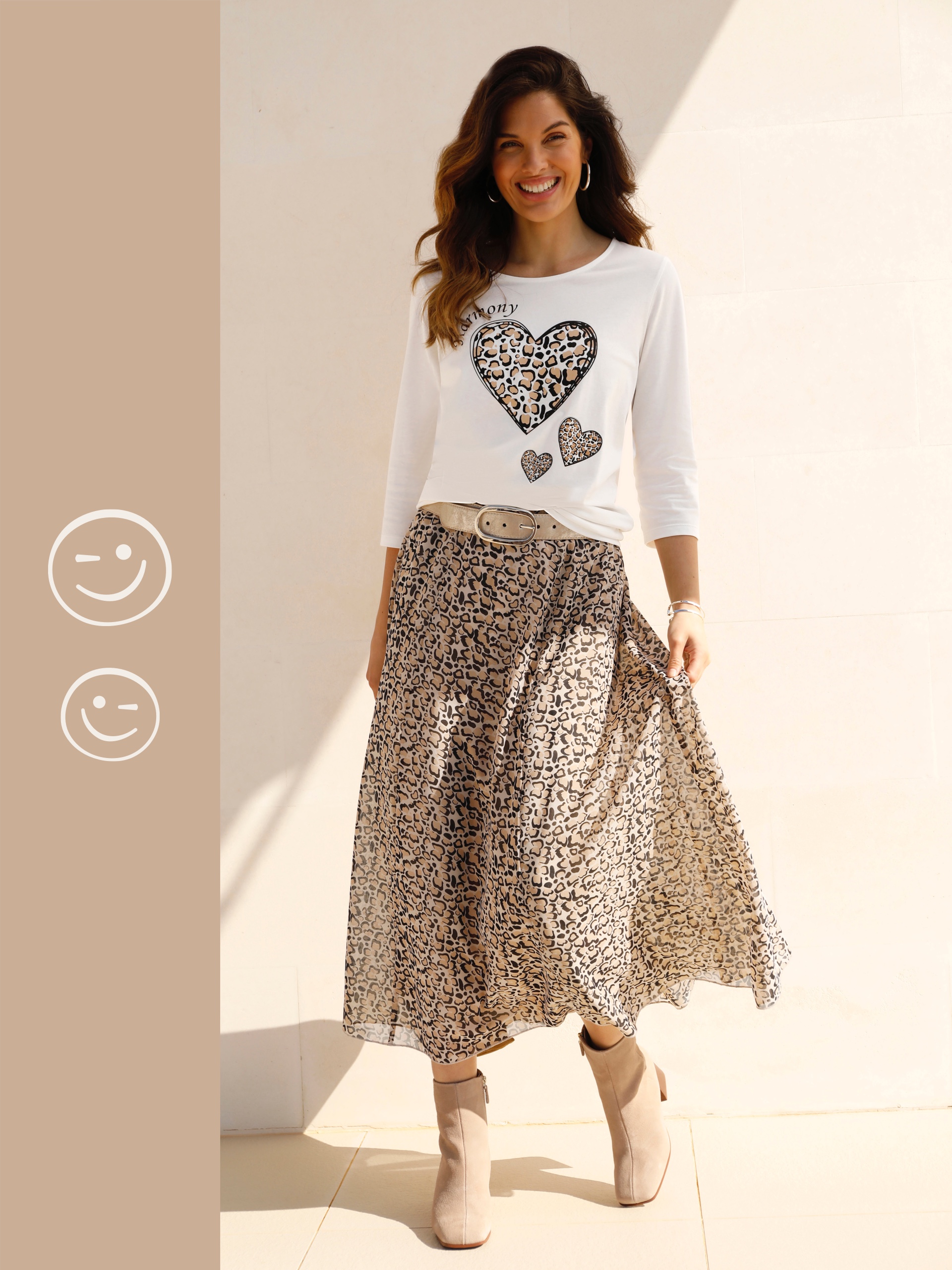 Ein Model trägt ein weißes Oberteil mit Herzmotiv und einen langen, gemusterten Rock. Die Schuhe sind beige. Der Hintergrund ist hell.