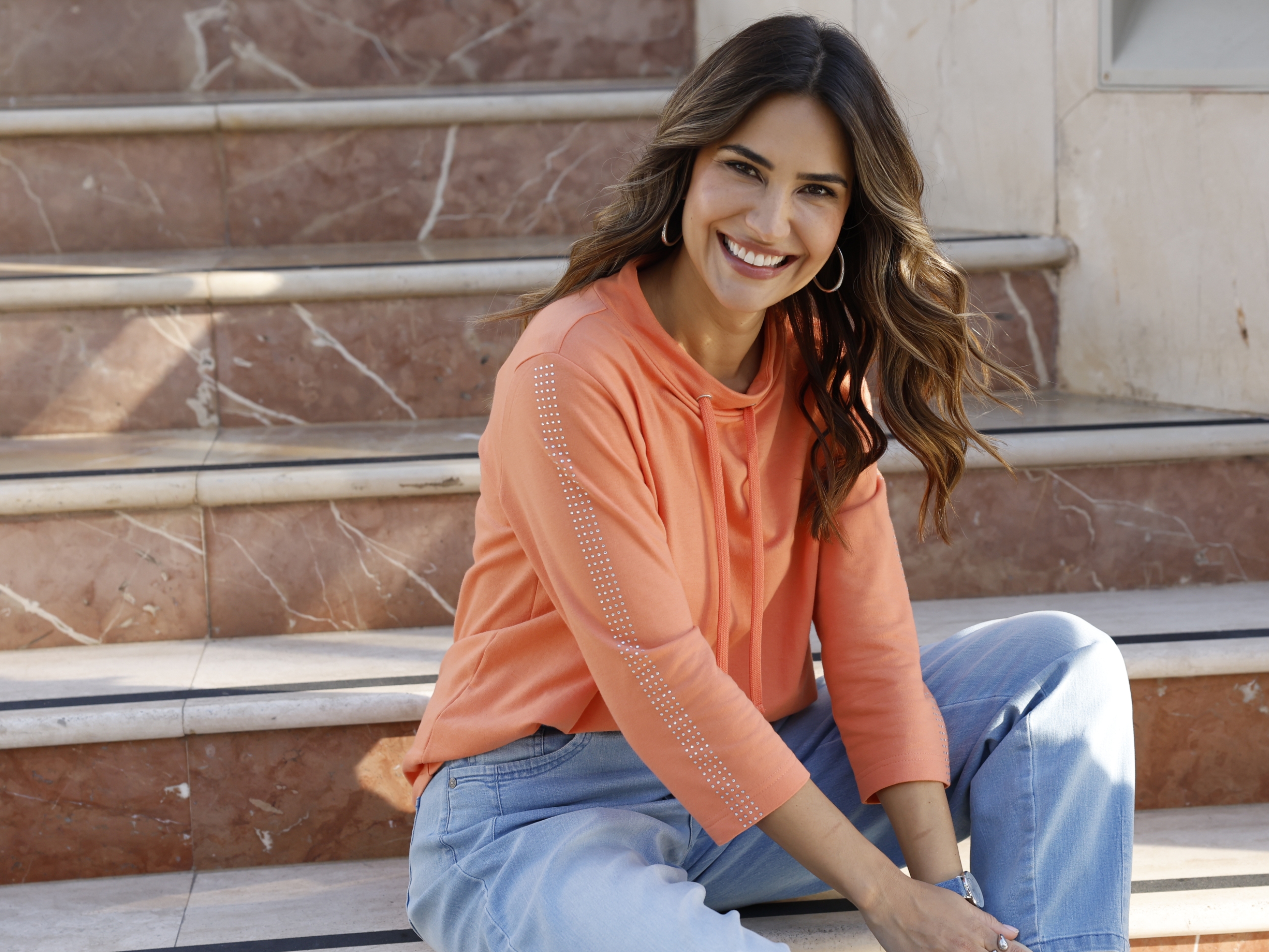 Het model draagt een oranje top met driekwart mouwen en lichte spijkerbroek voor een lichte achtergrond.