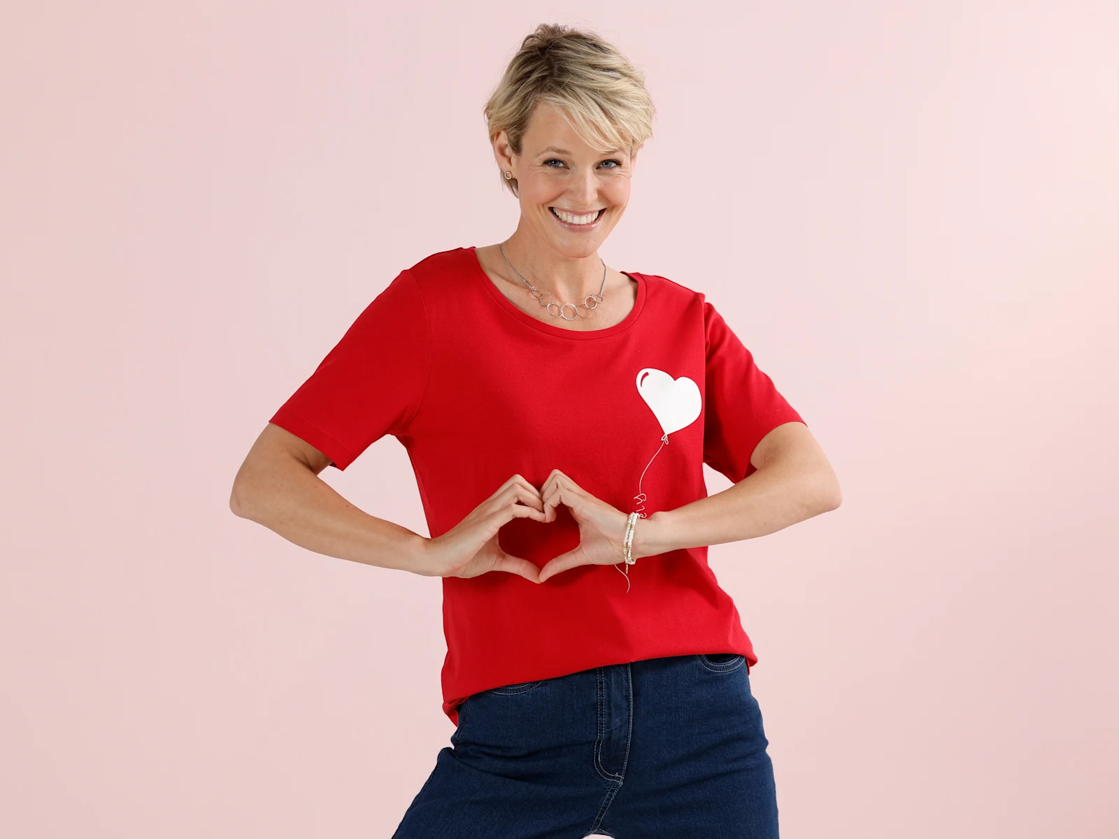 Ein Model mit kurzen blonden Haaren trägt ein rotes T-Shirt mit weißem Herzballon-Motiv und formt mit den Händen ein Herz vor einem rosa Hintergrund.