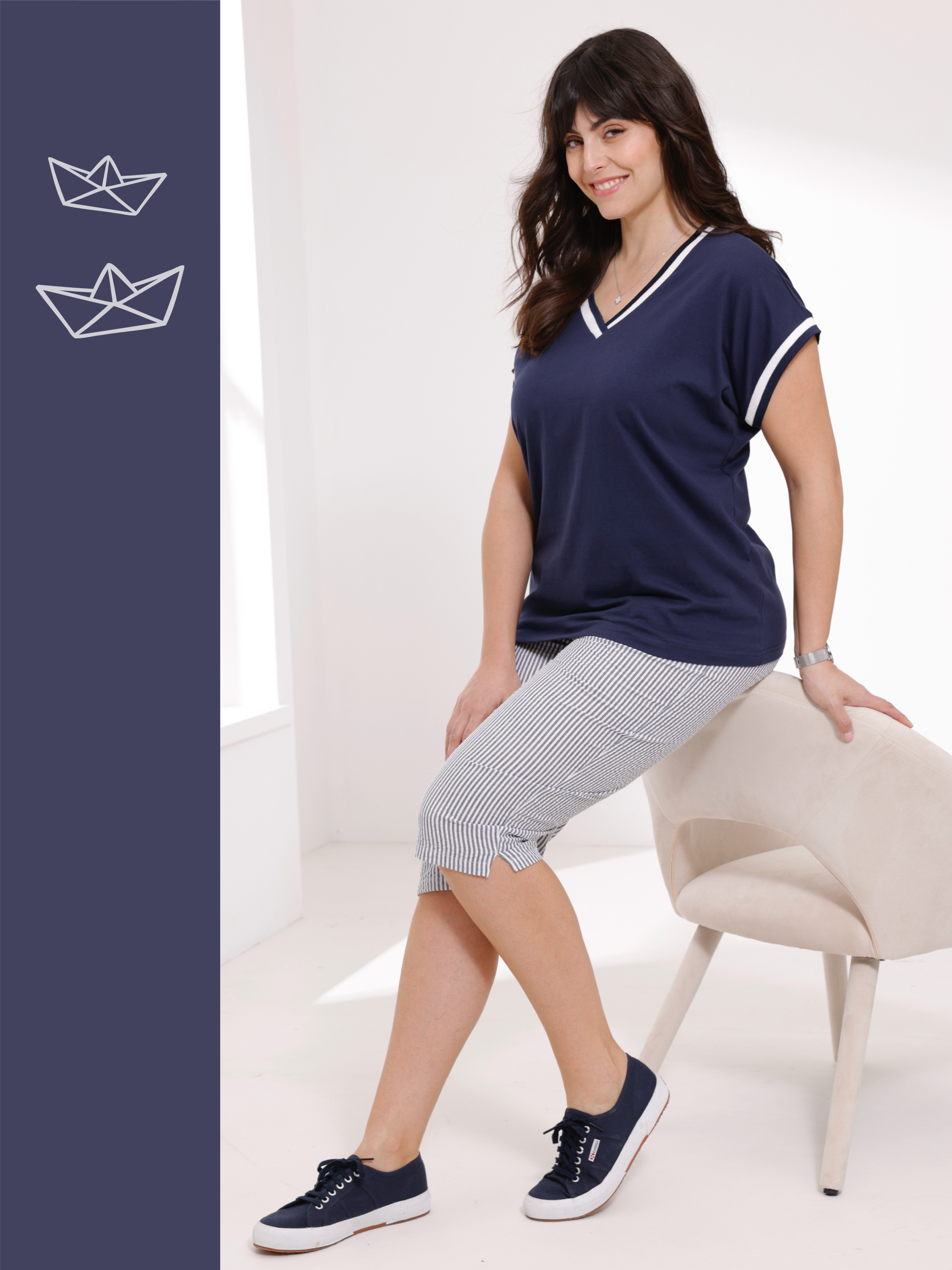 Vrouw draagt een donkerblauw shirt met V-hals en witte strepen, gestreepte driekwartbroek en blauwe sneakers, zittend op een beige stoel.