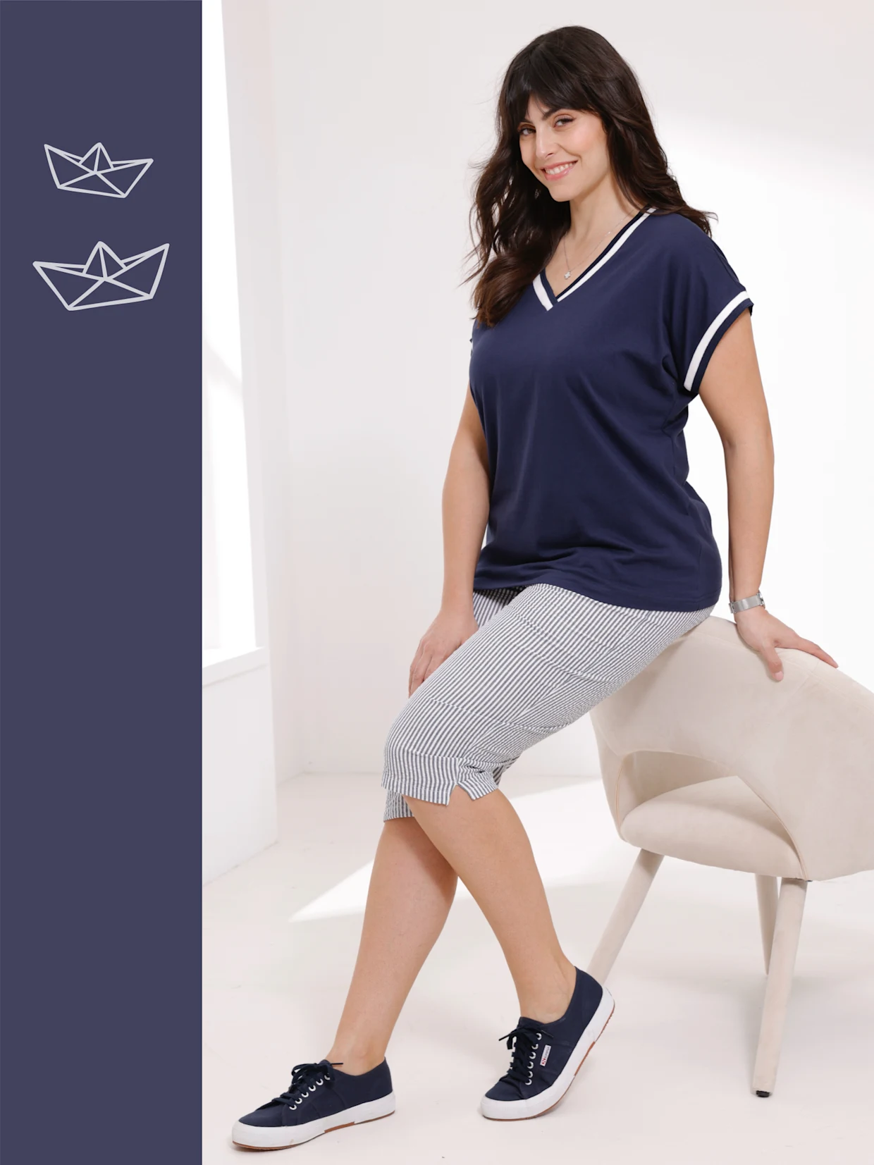 Žena sedící na béžové židli, oblečená v tmavě modrém tričku s bílými detaily, pruhovaných kalhotách a tmavě modrých teniskách. Žena sedící na béžové židli, oblečená v tmavě modrém tričku s bílými detaily, pruhovaných kalhotách a tmavě modrých teniskách.