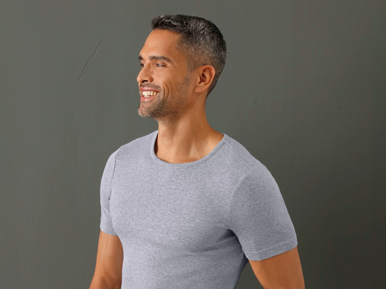 Een model met kort donker haar draagt een grijs T-shirt en lacht, voor een donkere achtergrond.