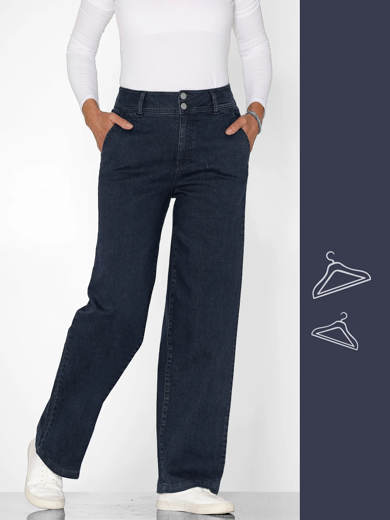 Ein Model trägt eine weiße Langarmbluse und dunkelblaue Jeans, mit den Händen in den Taschen, vor neutralem Hintergrund.