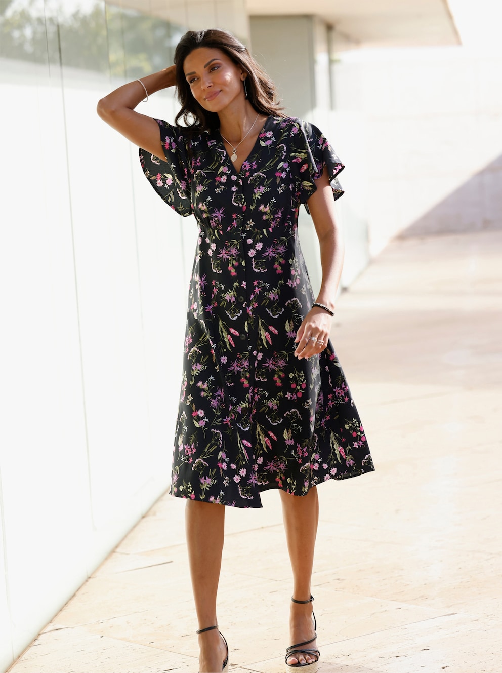 Frau mit Blumendruckkleid und scharzen Keilriemensandaletten. Das Kleid hat einen V-Ausschnitt und  kurze Flügel-Ärmel.