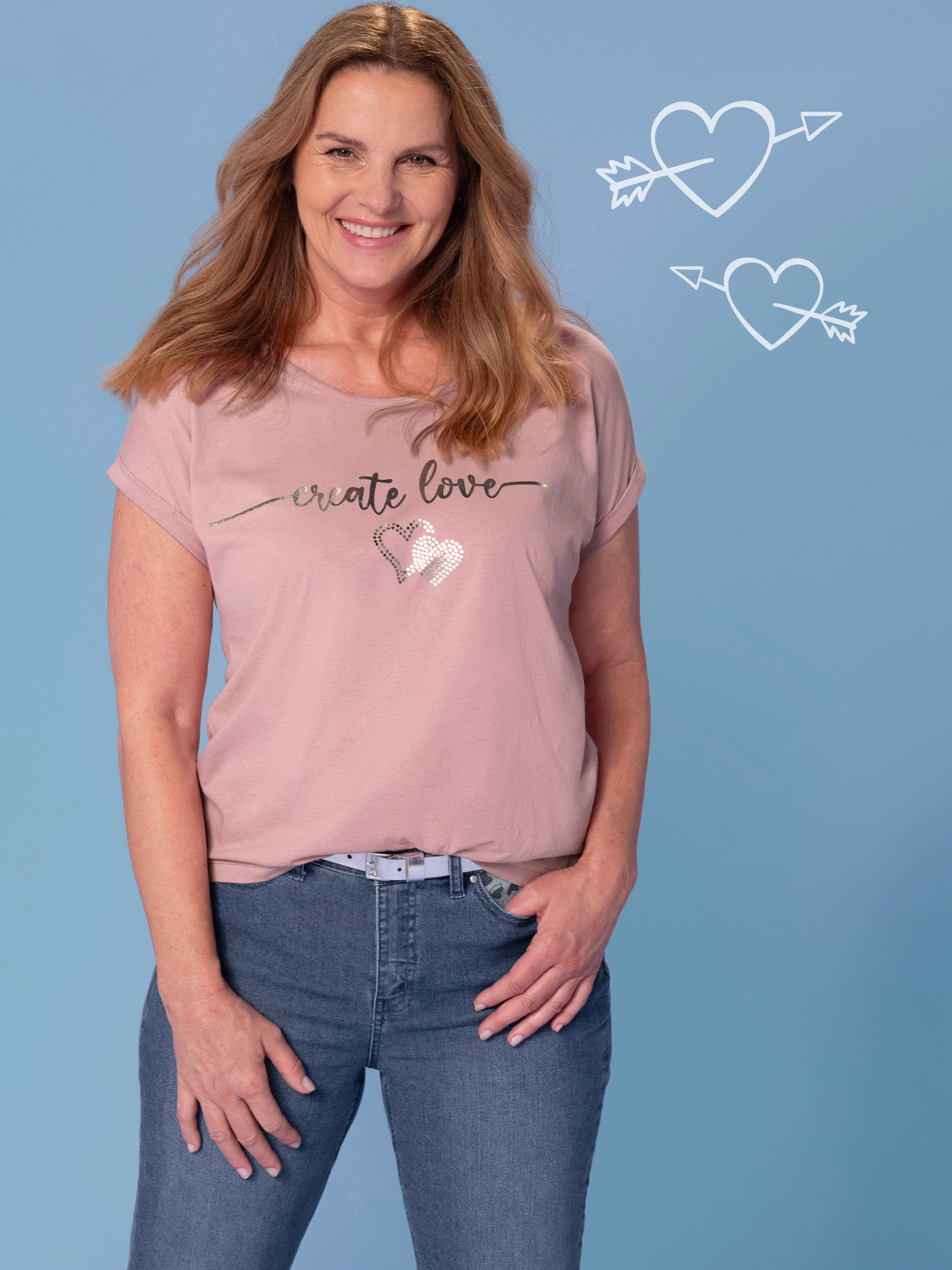 Frau trägt ein rosafarbenes T-Shirt mit der Aufschrift 'create love' und zwei Herzen aus Pailletten, kombiniert mit einer blauen Jeans vor einem hellblauen Hintergrund.