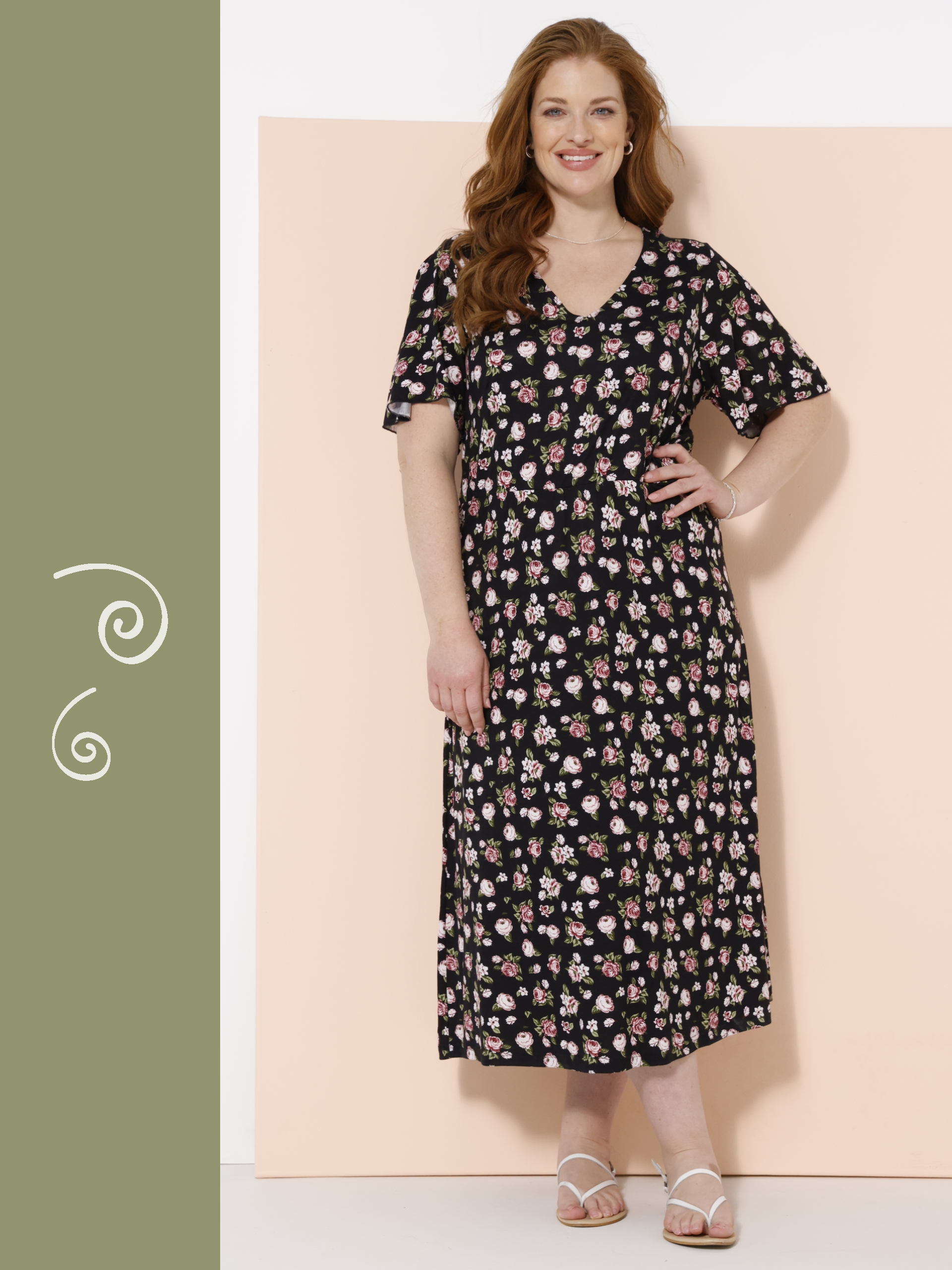 Ein Model steht vor einer zweifarbigen Wand, trägt ein schwarzes, knöchellanges Kleid mit kurzen Ärmeln und floralem Muster sowie weiße Sandalen.