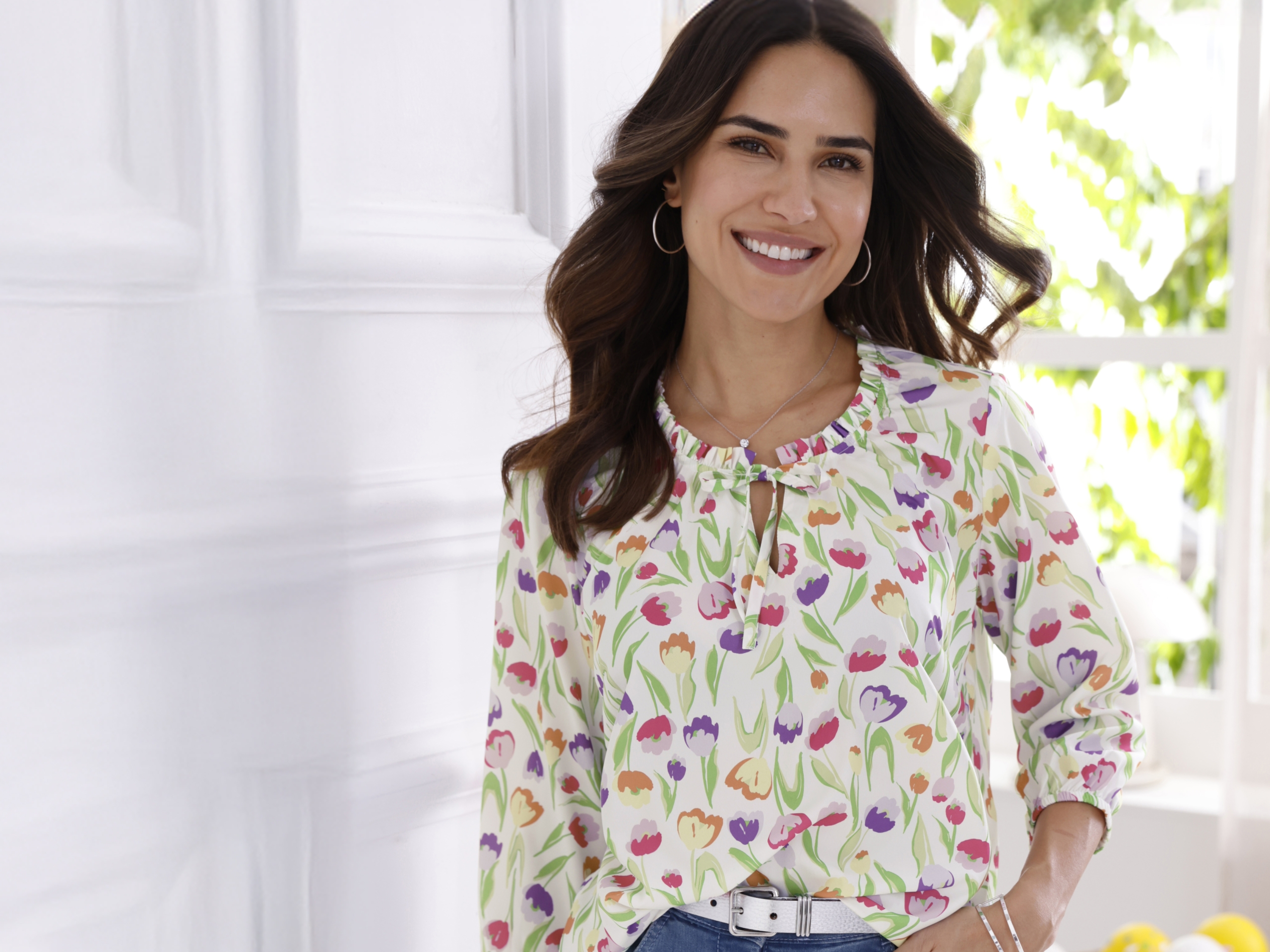 Modelka má na sebe bielu blúzku s farebným vzorom tulipánov a 3/4 rukávmi pred svetlým pozadím.