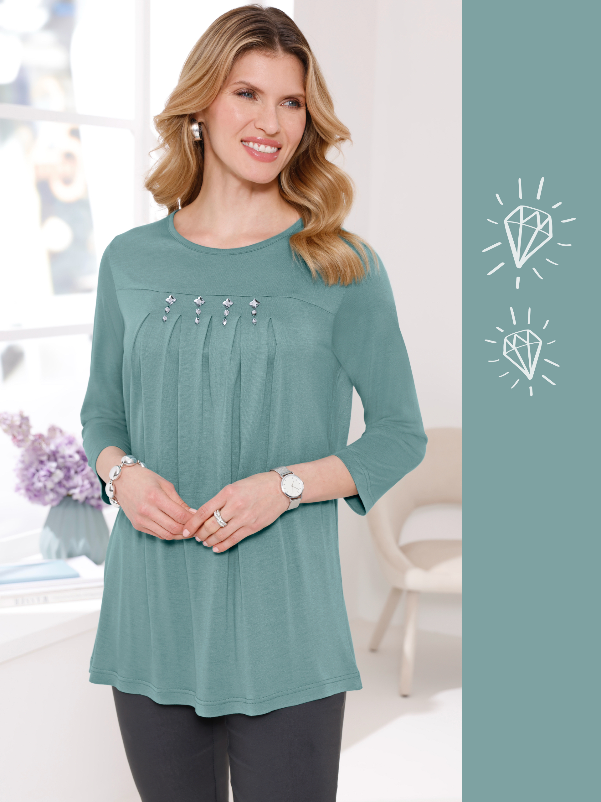 En modell med blont hår bär en grön blus med smyckesstenar, armband, ring och en silverfärgad klocka. Hon ler.