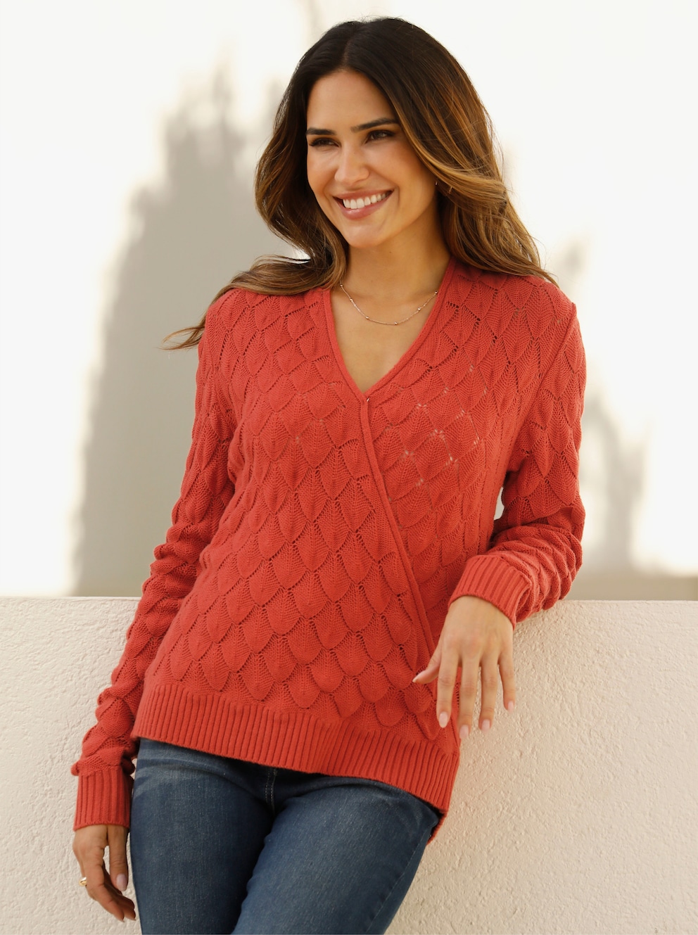 Weibliches Model stehend vor einer Wand. Das Model trägt eine blaue Jeans und einen Ajourpullover in Wickel-Optik in der Farbe terra.