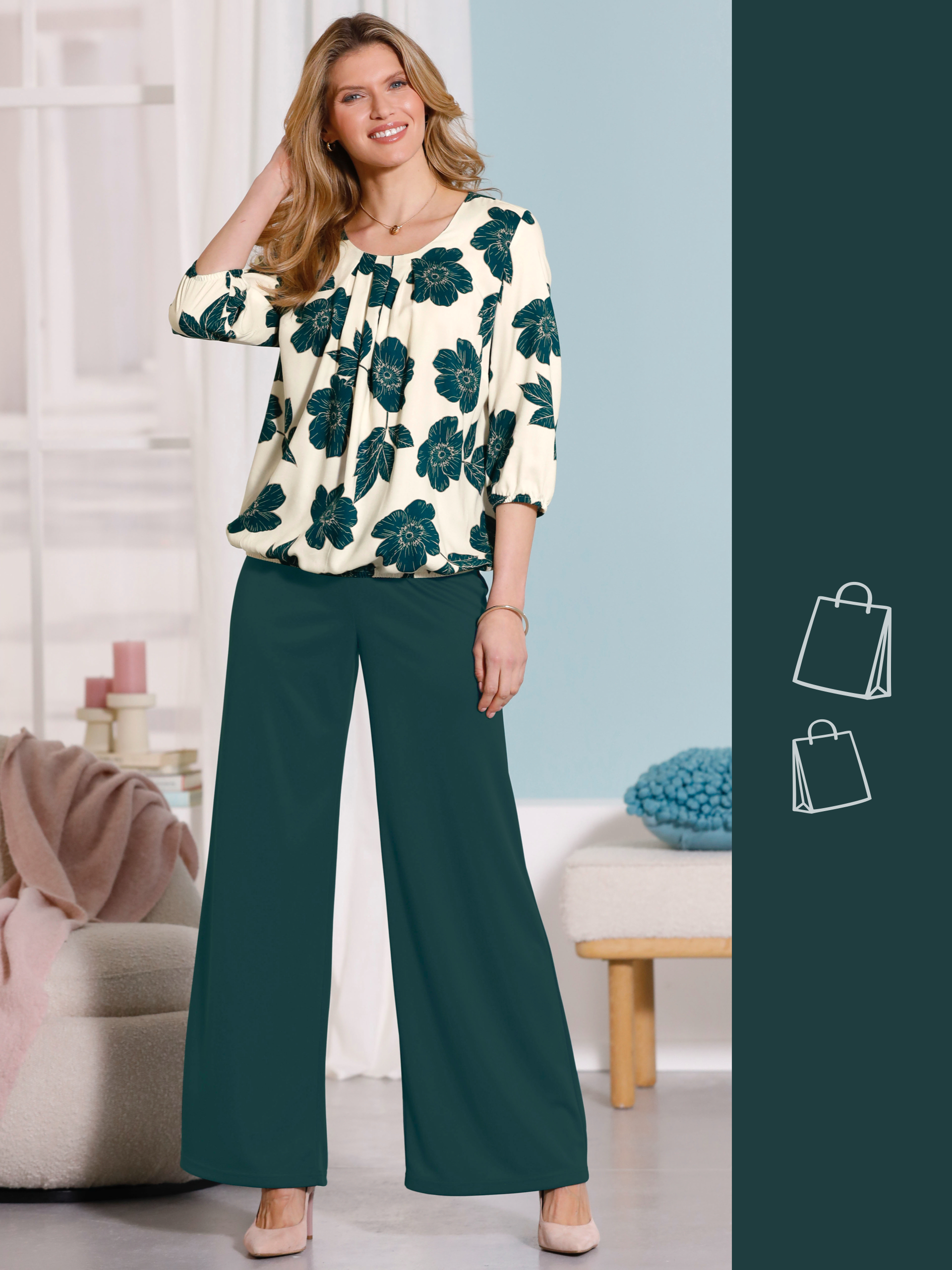 Ein Model trägt eine weiße Bluse mit großen grünen Blumen und weite grüne Hosen, steht in einem hellen Wohnzimmer.