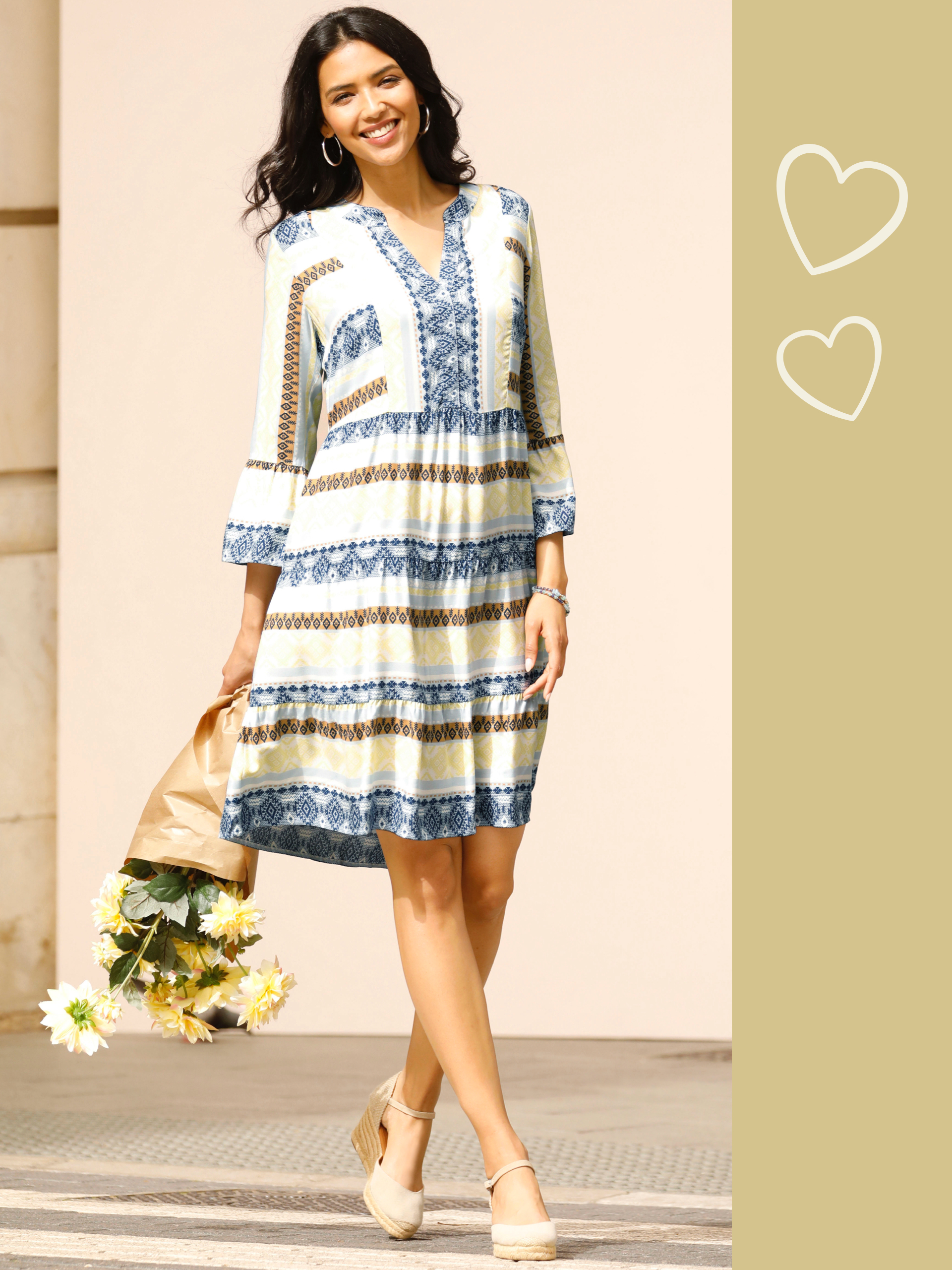 Ein Model trägt ein knielanges, gemustertes Kleid mit 3/4-Ärmeln und V-Ausschnitt, dazu bequeme beige Schuhe und hält einen Strauß Blumen.