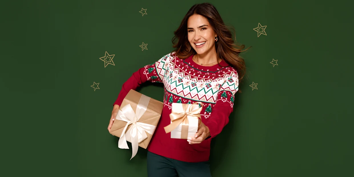 Ein Model lächelt mit Geschenken in den Händen. Sie trägt einen roten gemusterten Weihnachtspullover.