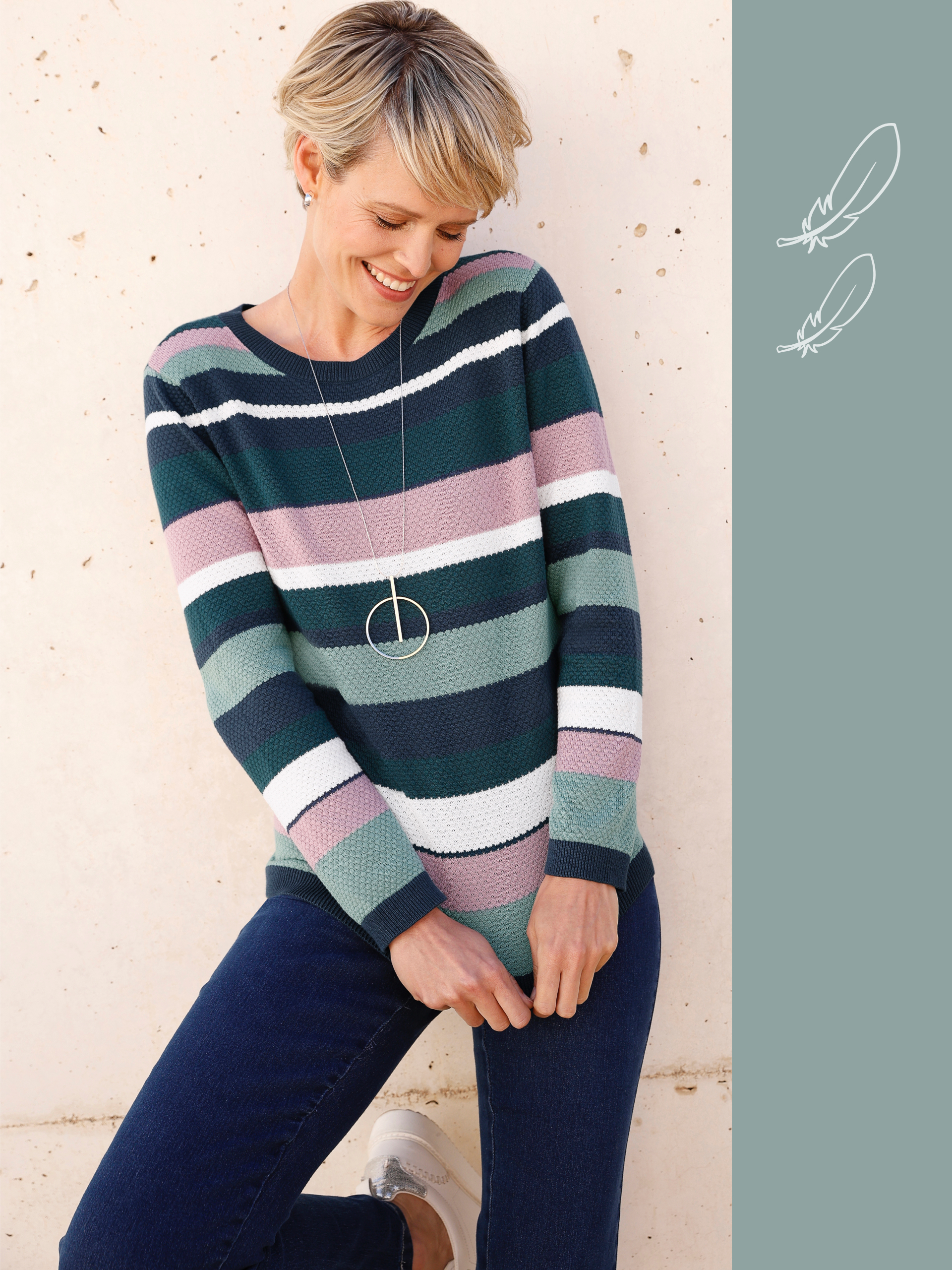 Model má na sebe pruhovaný sveter v zelených, bielych a ružových tónoch, modré džínsy a dlhý strieborný náhrdelník.