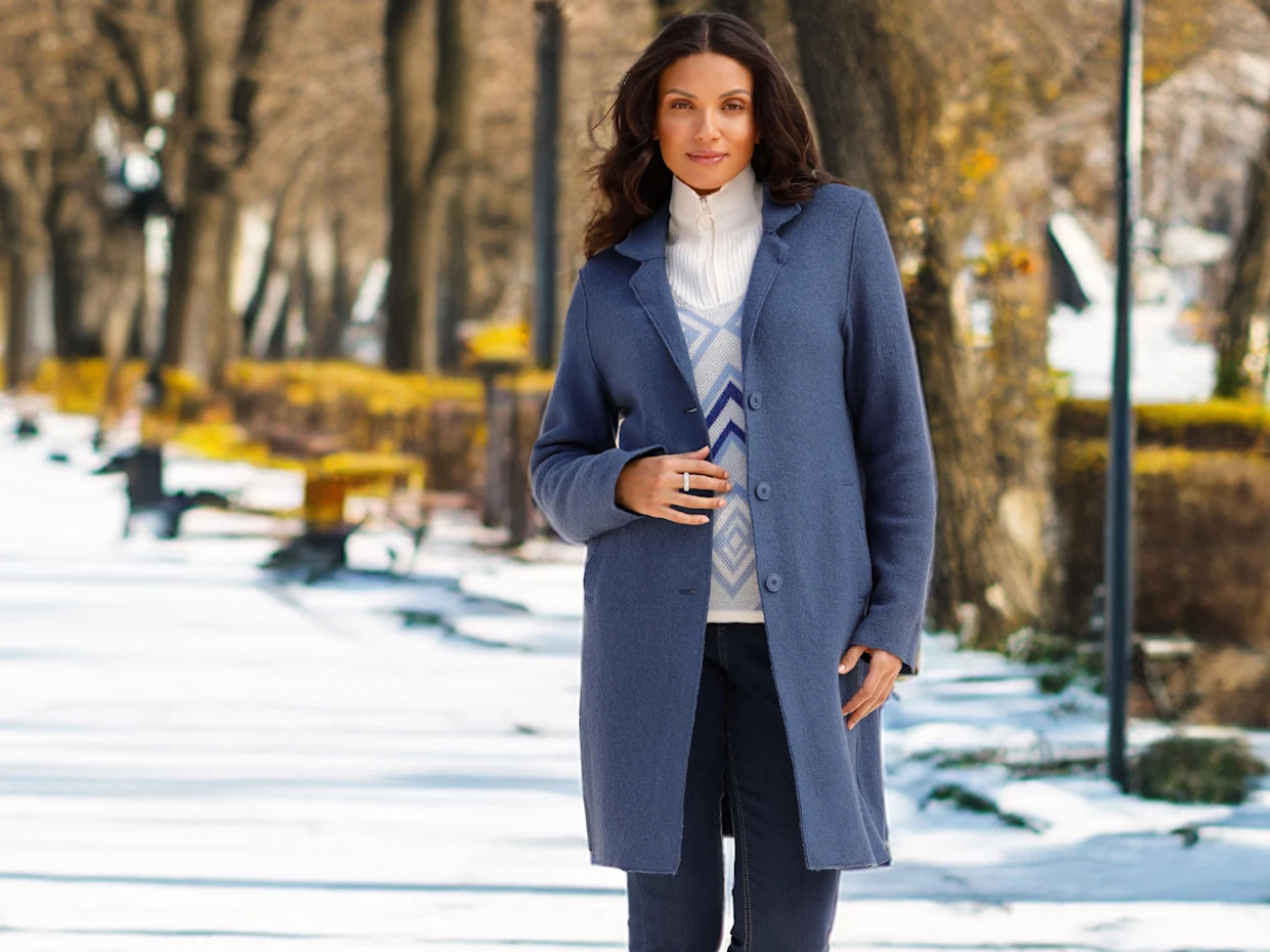 Ein Model mit dunklen Haaren trägt einen langen blauen Mantel, eine weiße Strickjacke und Jeans.