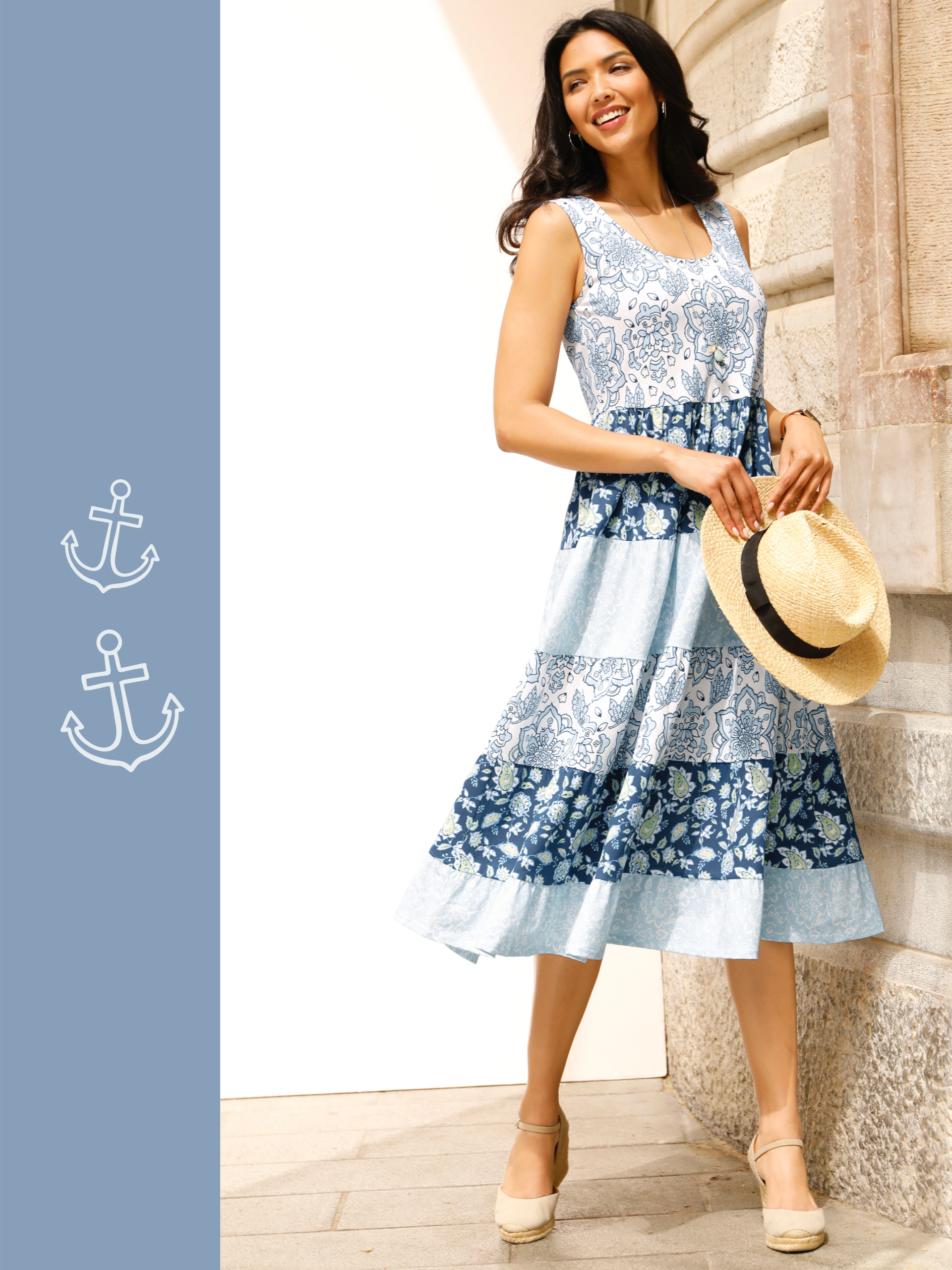 Ein Model trägt ein ärmelloses, midilanges Kleid mit blau-weißem Blumenmuster und hält einen Strohhut.