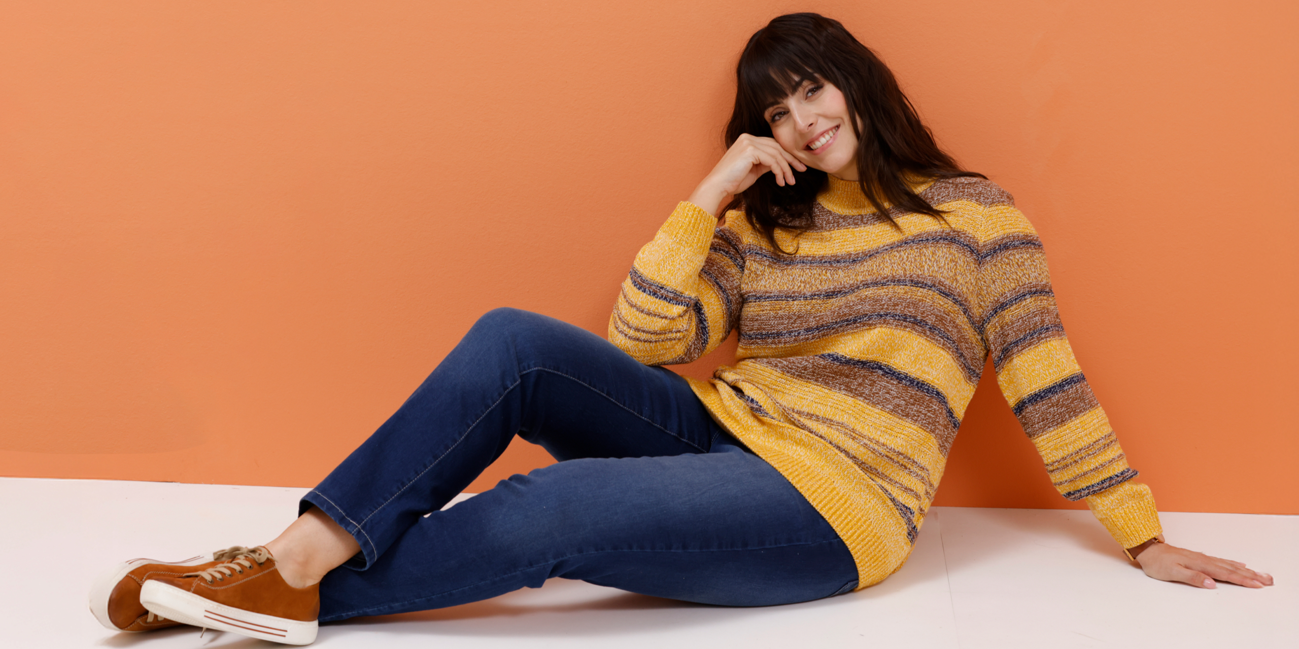 Ein Model trägt einen gestreiften Pullover in Gelb- und Brauntönen und blaue Jeans.
