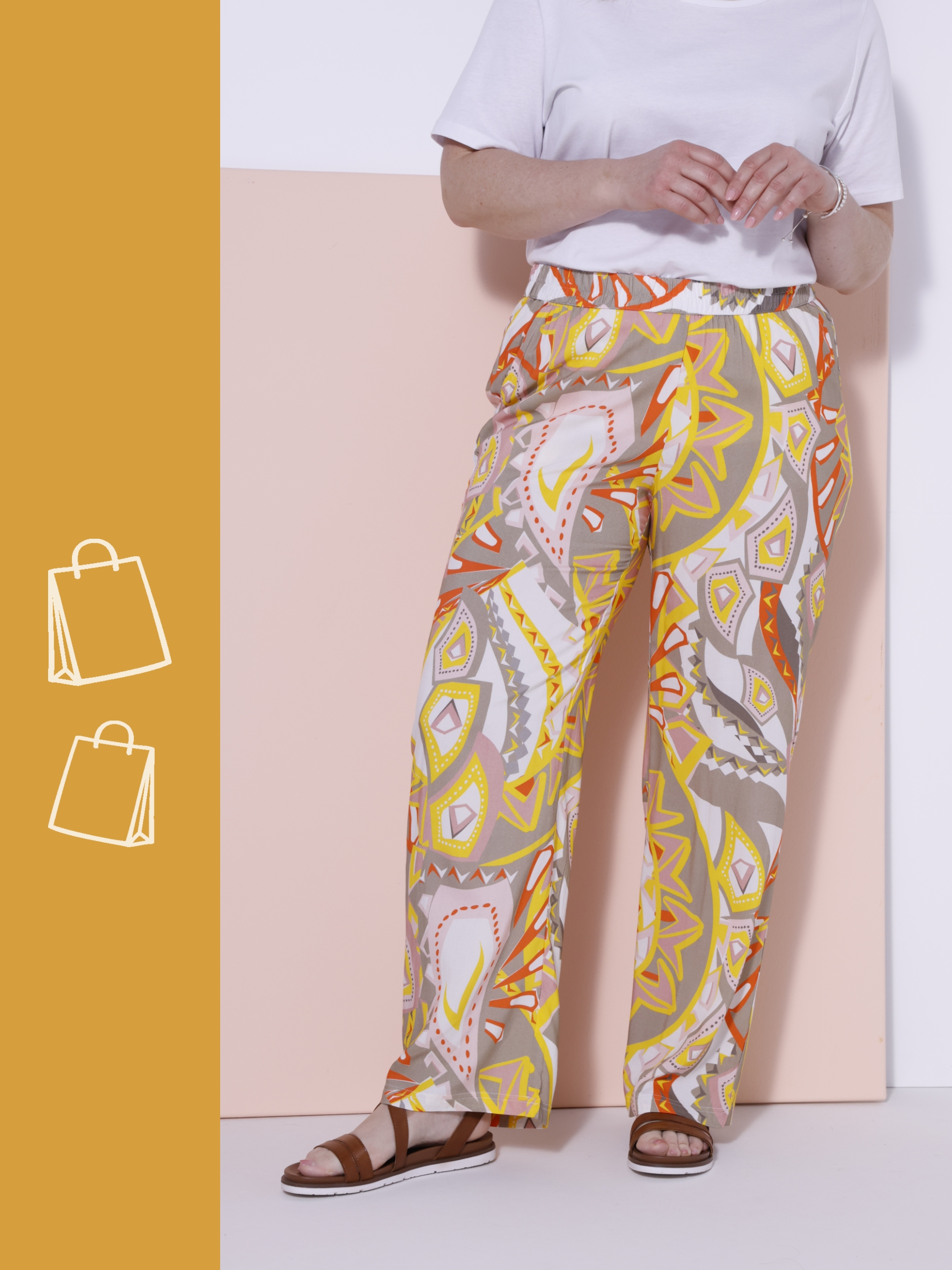 Ein Model steht vor einer zweifarbigen Wand, trägt eine weiße Kurzarmbluse und bunte, weit geschnittene Hosen mit grafischem Muster sowie braune Sandalen.