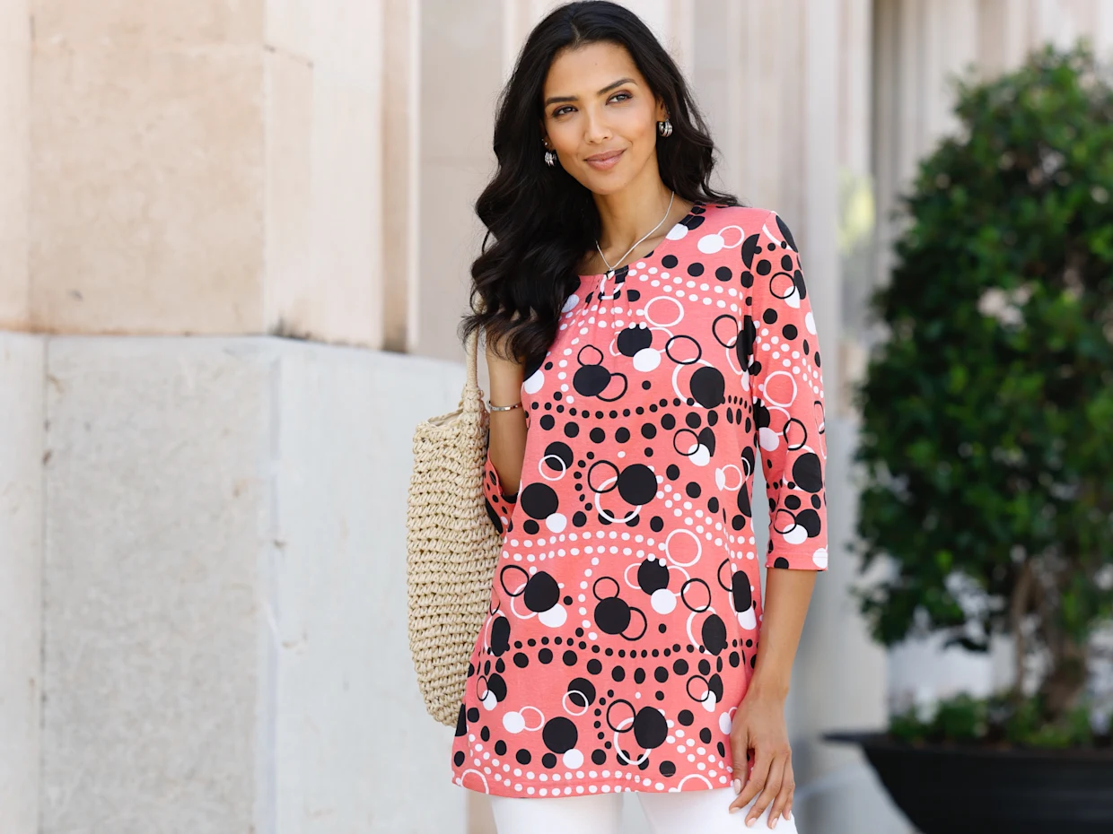 Model má na sobě růžové puntíkované tričko s 3/4 rukávy a drží pletenou tašku.