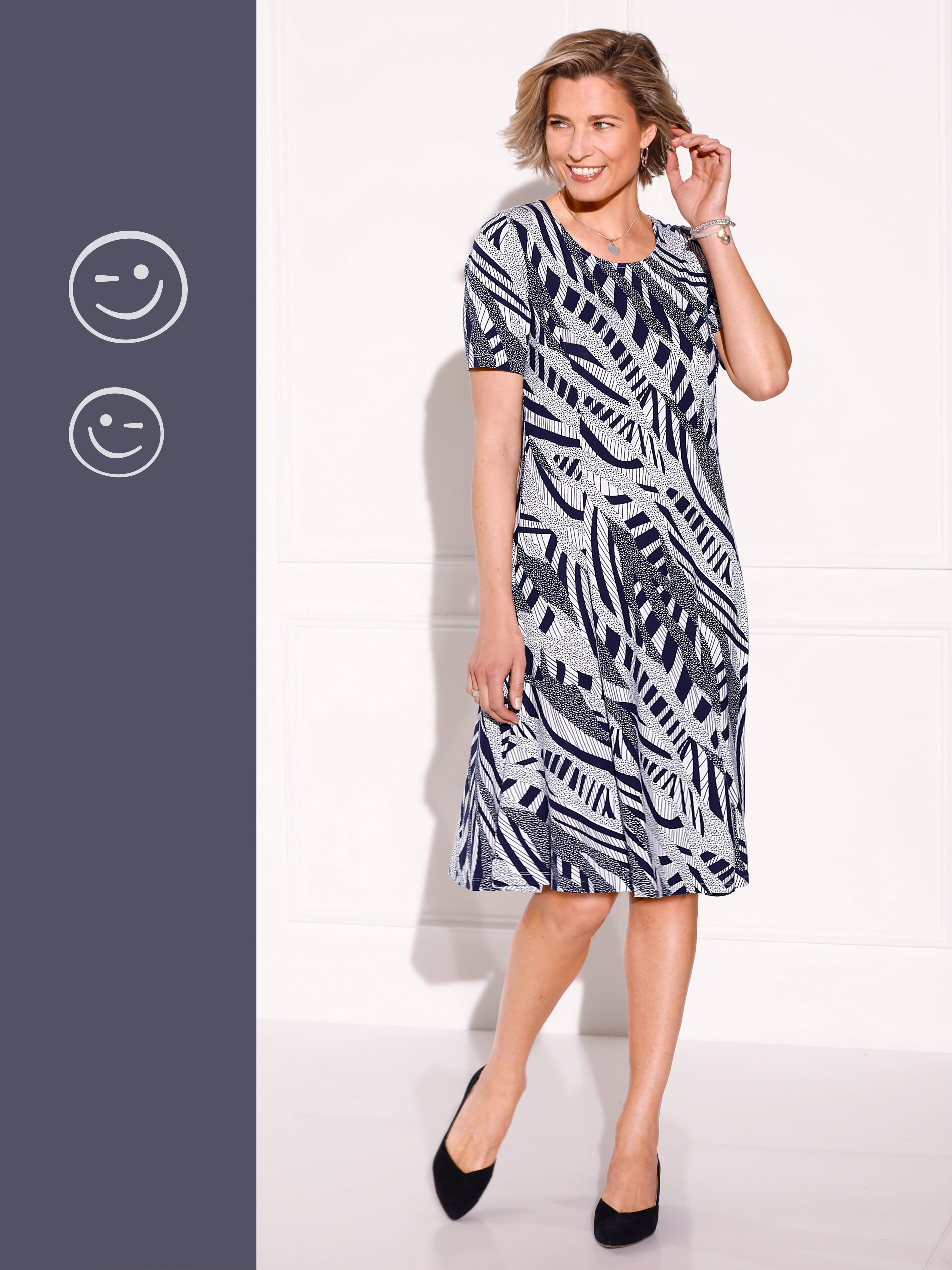 Ein Model trägt ein knielanges Kleid mit schwarz-weißem Blattmuster und schwarzen Schuhen.