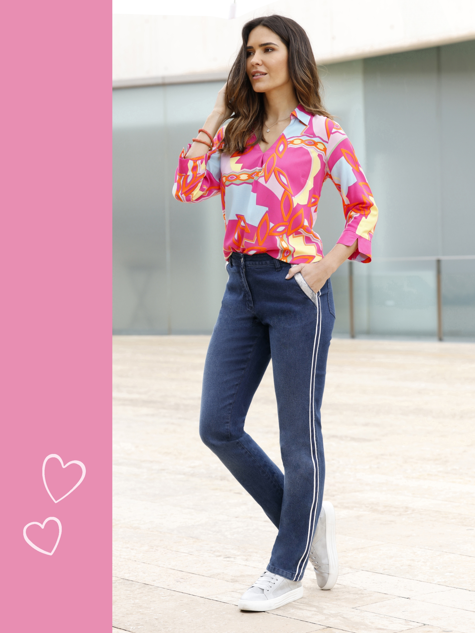 Een model draagt een kleurrijke blouse met roze en oranje patroon, gecombineerd met blauwe spijkerbroeken met witte strepen aan de zijkant en witte sneakers.