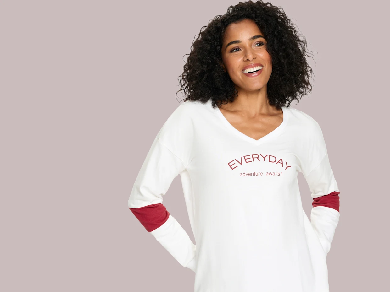 Modelka má na sebe biele tričko s dlhým rukávom a výstrihom do V s červeným nápisom 'EVERYDAY adventure awaits!' a červenými záplatami na lakťoch.