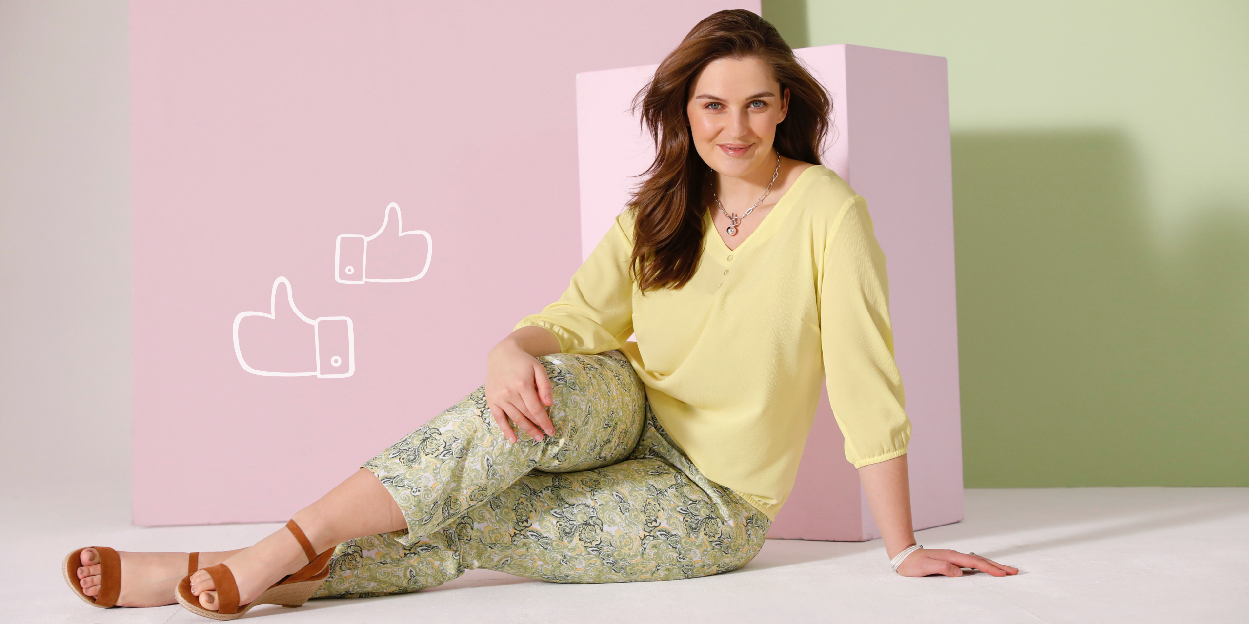 Ein Model sitzt auf dem Boden, trägt eine gelbe Bluse, gemusterte Hose und braune Sandalen, vor pastellfarbenem Hintergrund.