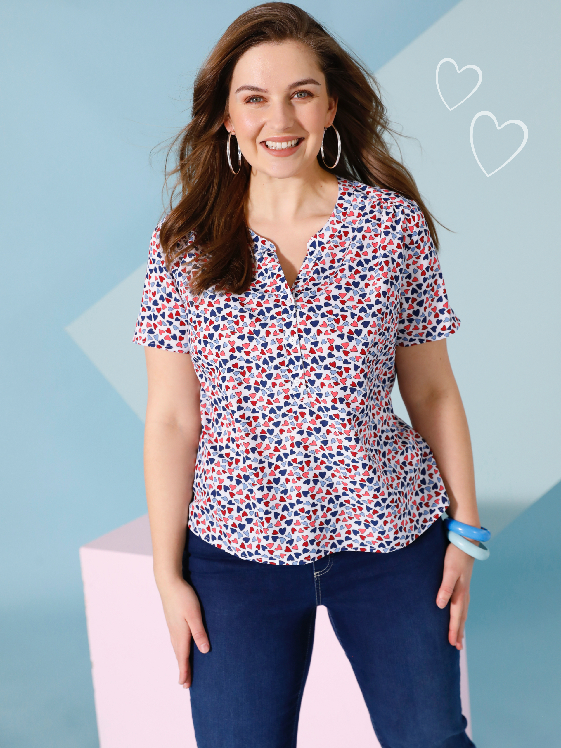Ein Model trägt eine kurzärmelige Bluse mit Herzmuster und dunkle Jeans vor pastellfarbenem Hintergrund.