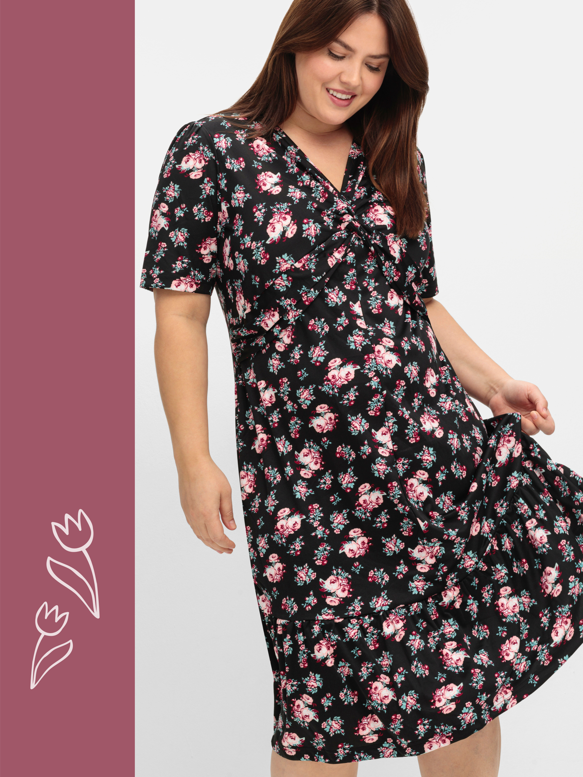 Ein Model trägt ein schwarzes Kleid mit kurzem Arm und rosa-weißem Blumenmuster vor weißem Hintergrund.