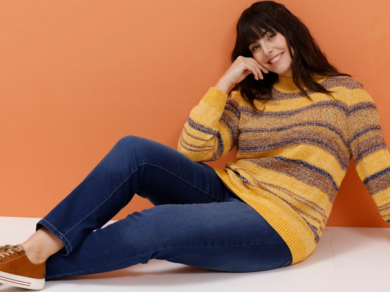 Ein Model trägt einen gestreiften Pullover in Gelb- und Brauntönen und blaue Jeans.