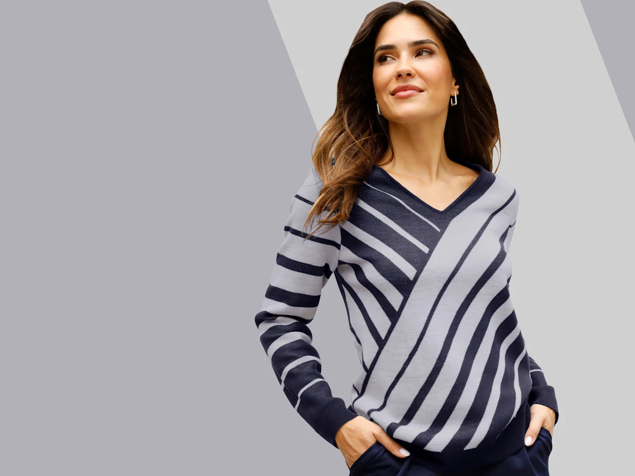 Model má na sebe sveter s výstrihom do V a diagonálnymi pruhmi v sivých a modrých odtieňoch a tmavomodré nohavice.