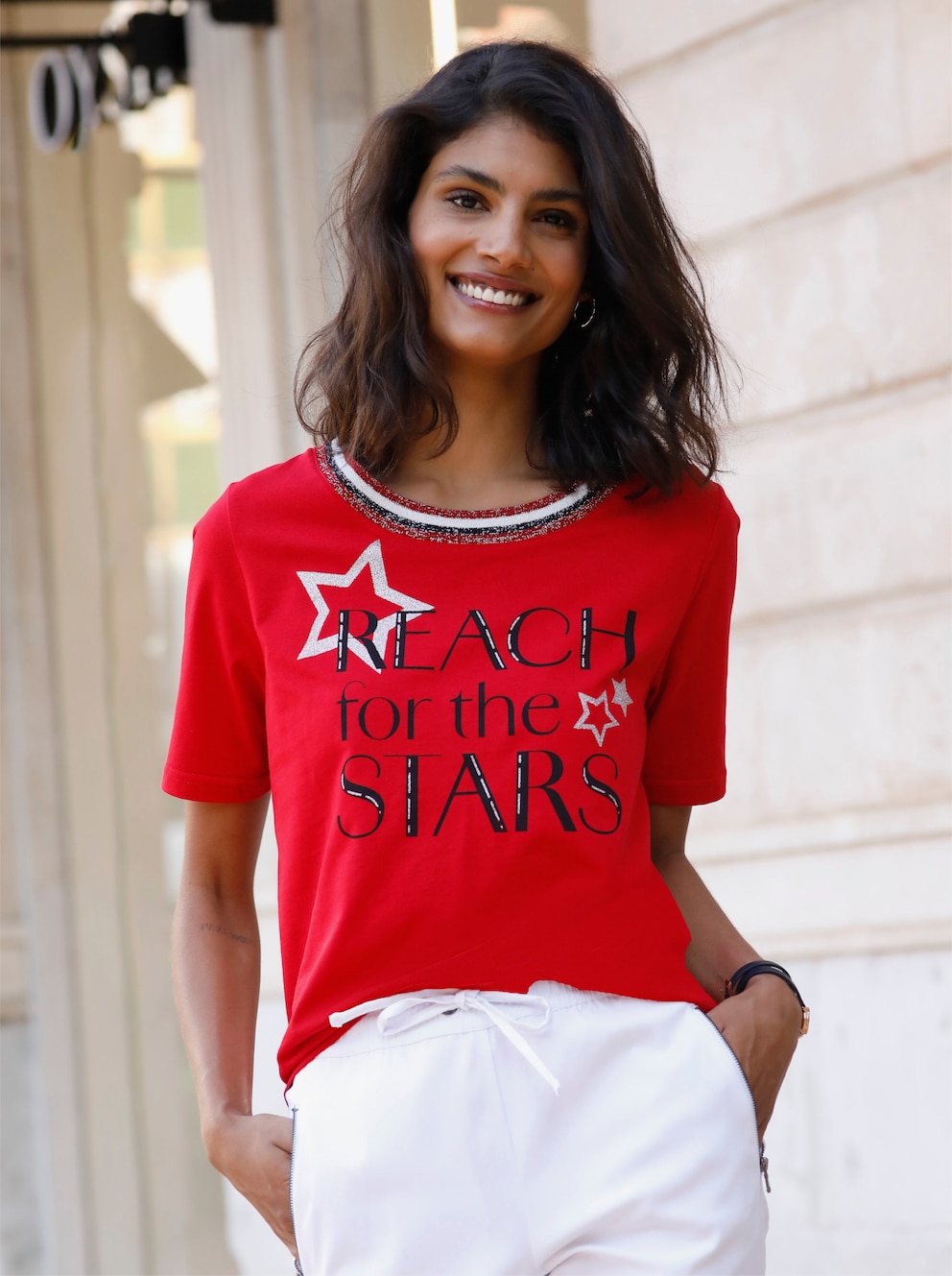 Frau stehend mit rotem Kurzarm-Shirt mit Statement-Spruch und Sternen in silberfarbig glitzerndem Druck auf der Vorderseite. Dazu trägt sie eine weiße Hose.